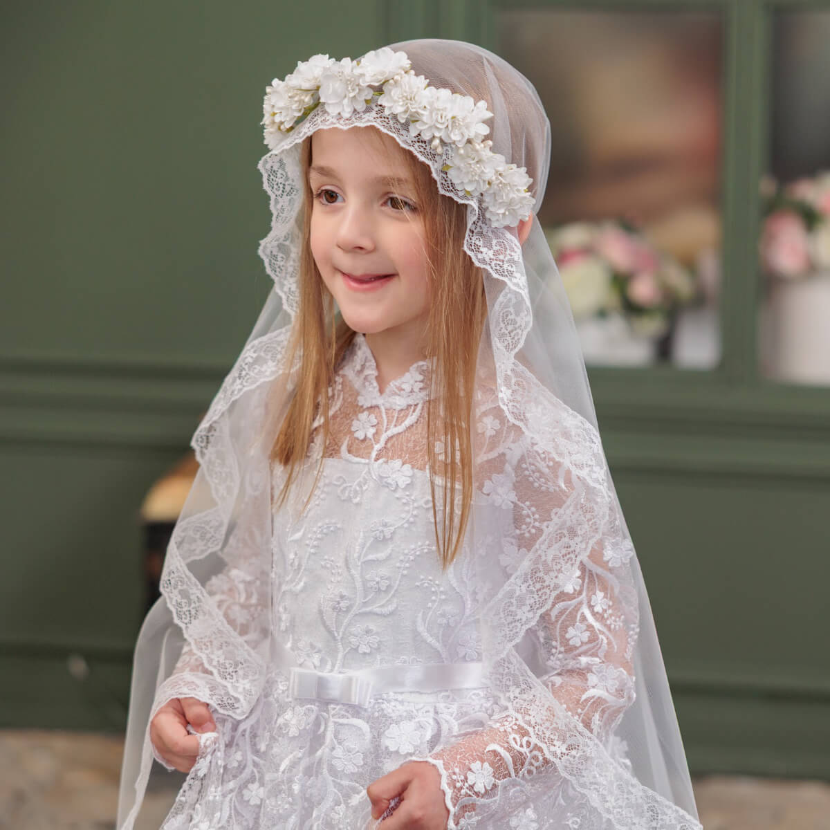 Young girl in a white lace communion dress with a veil and floral headband, standing outdoors.