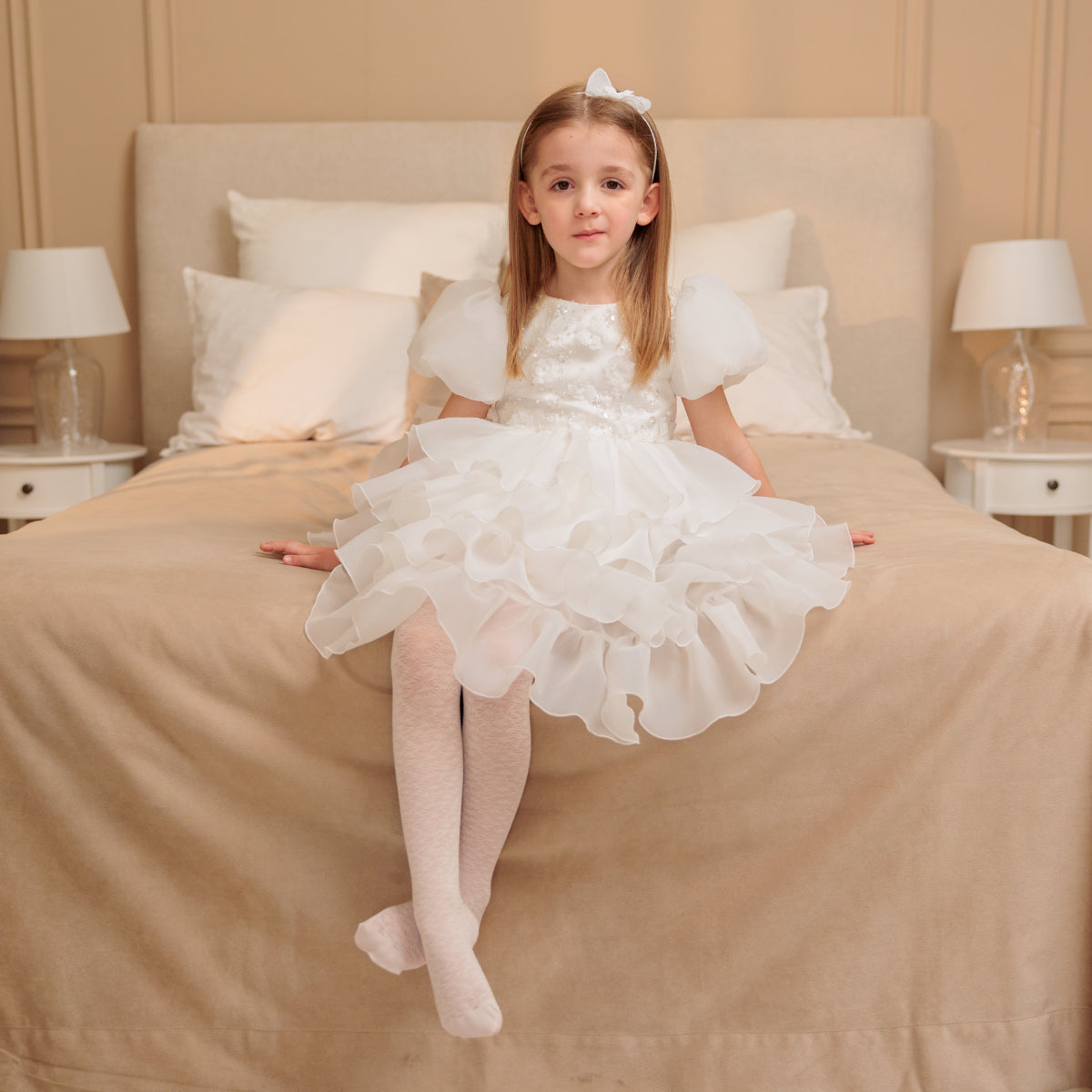 Young girl in a white dress sitting on a beige bed.