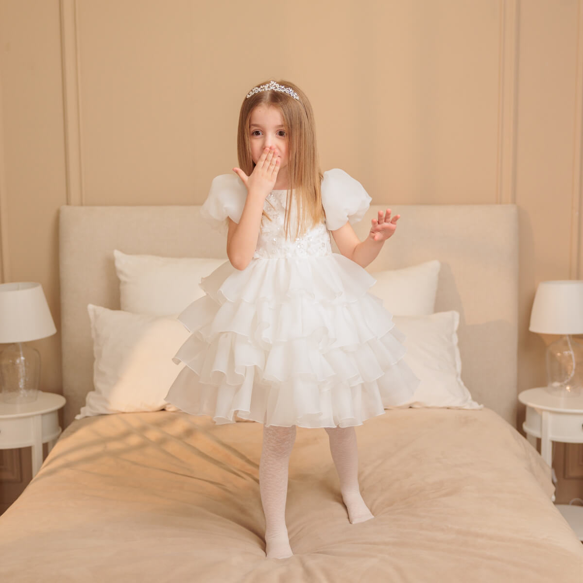 Young girl in a white dress standing on a bed in a bedroom setting