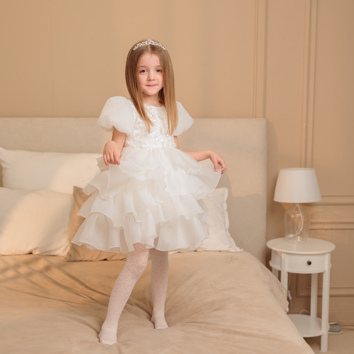 Young girl in a white ruffle flower girl dress standing on a bed in a bedroom setting