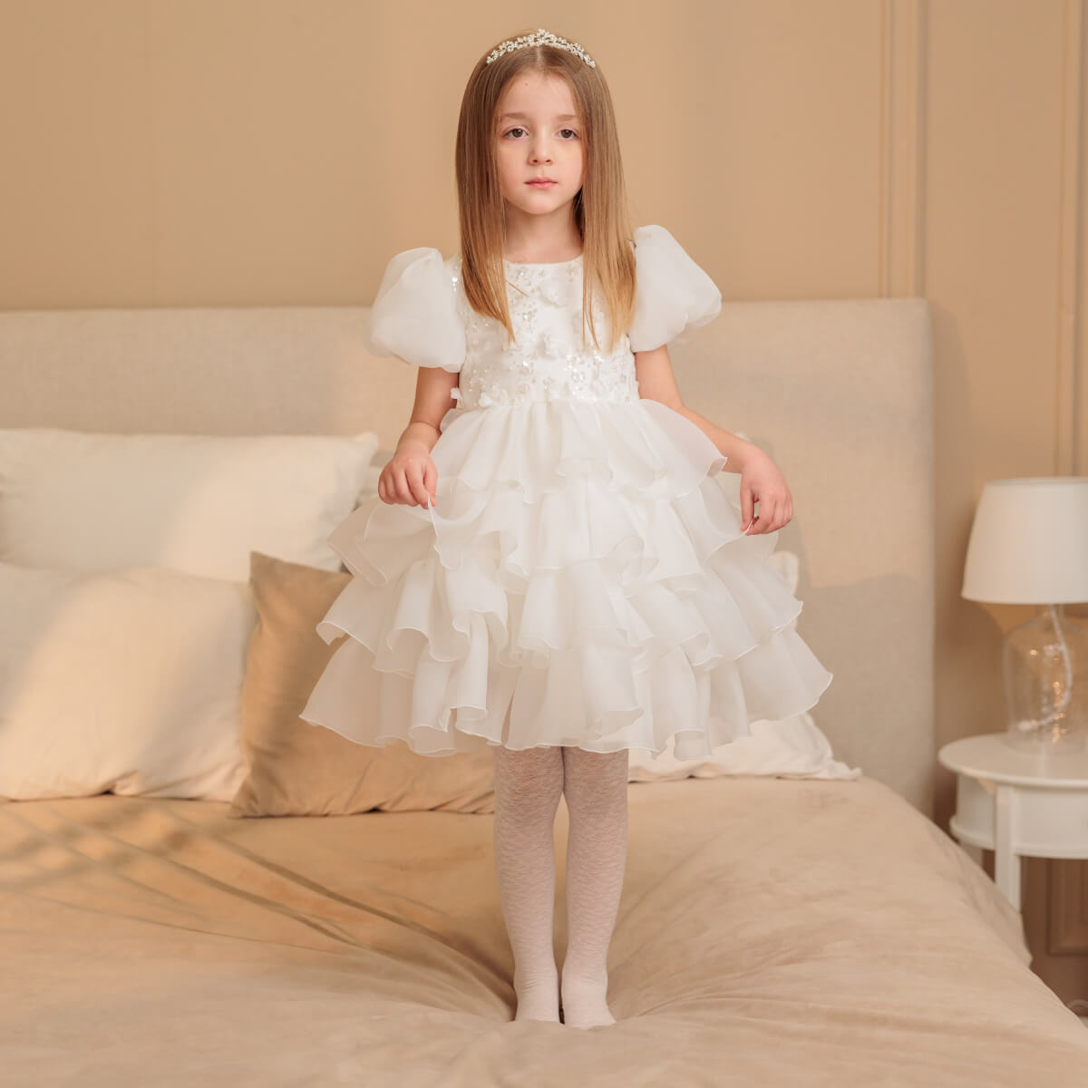 Young girl in a white dress standing on a bed in a softly lit room.