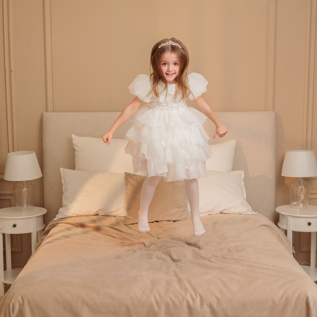 Young girl in a white dress jumping on a bed in a bedroom.