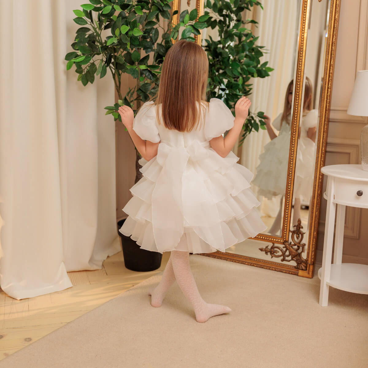 Child in a white dress standing in front of a large mirror with decorative plants.