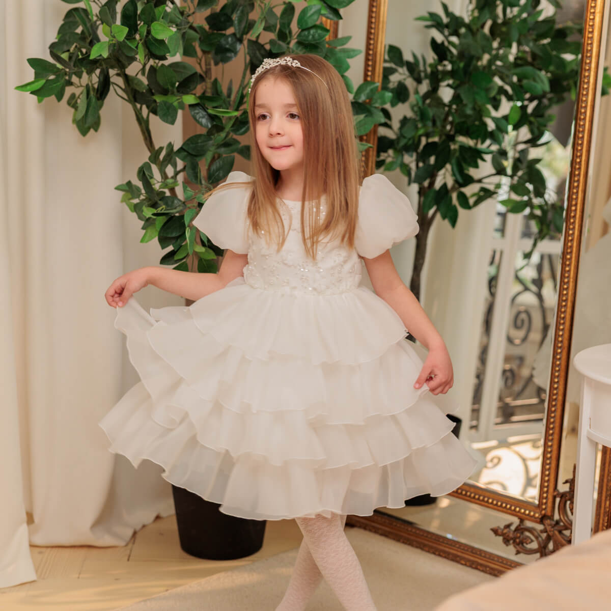 Young girl in a white dress standing in front of a mirror with decorative plants.