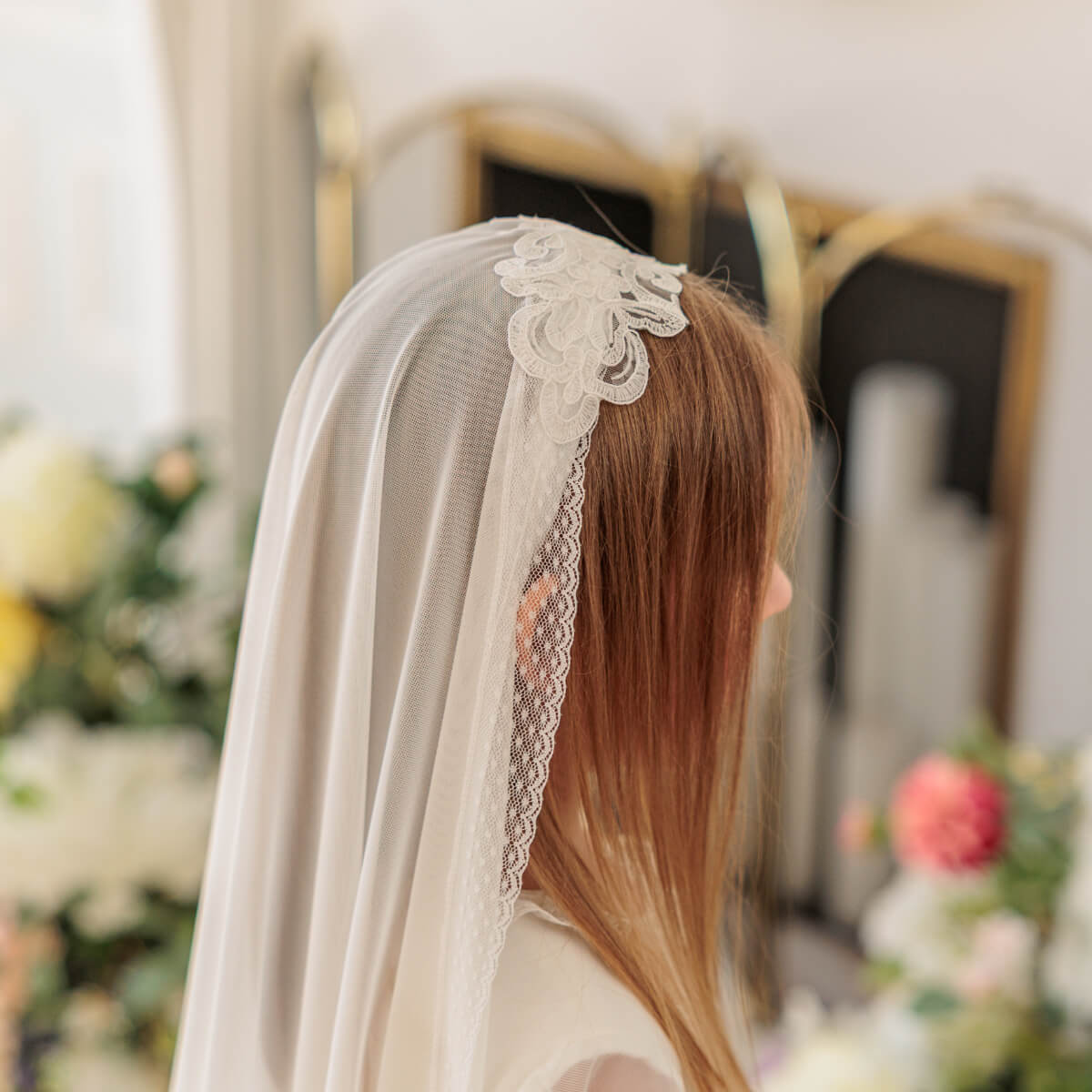 Person wearing a white veil with lace details, standing in a decorated indoor setting.