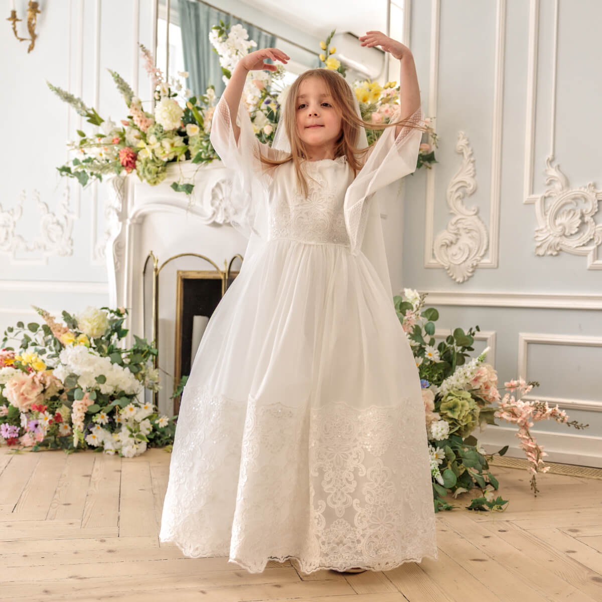 Young girl in Holy Communion Dress standing in a decorated room with floral arrangements.