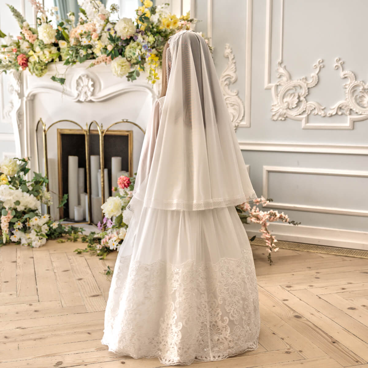 Person wearing a white lace communion dress with a veil in an elegant room with floral decorations.