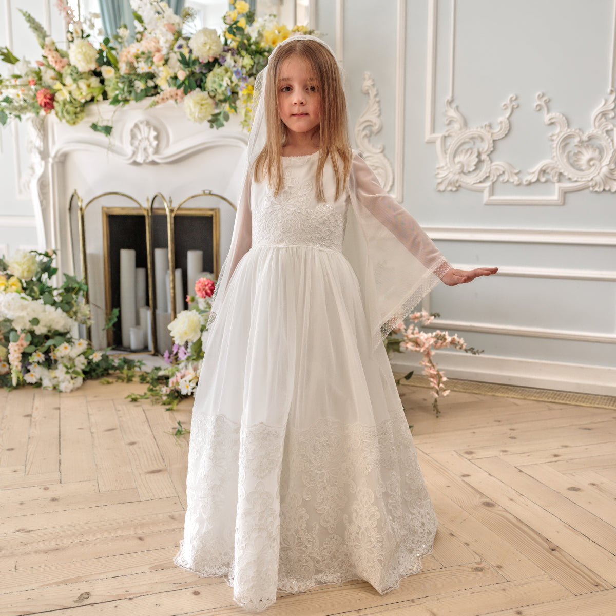 Young girl in a Holy Communion Dress standing in an elegant room with floral decorations.