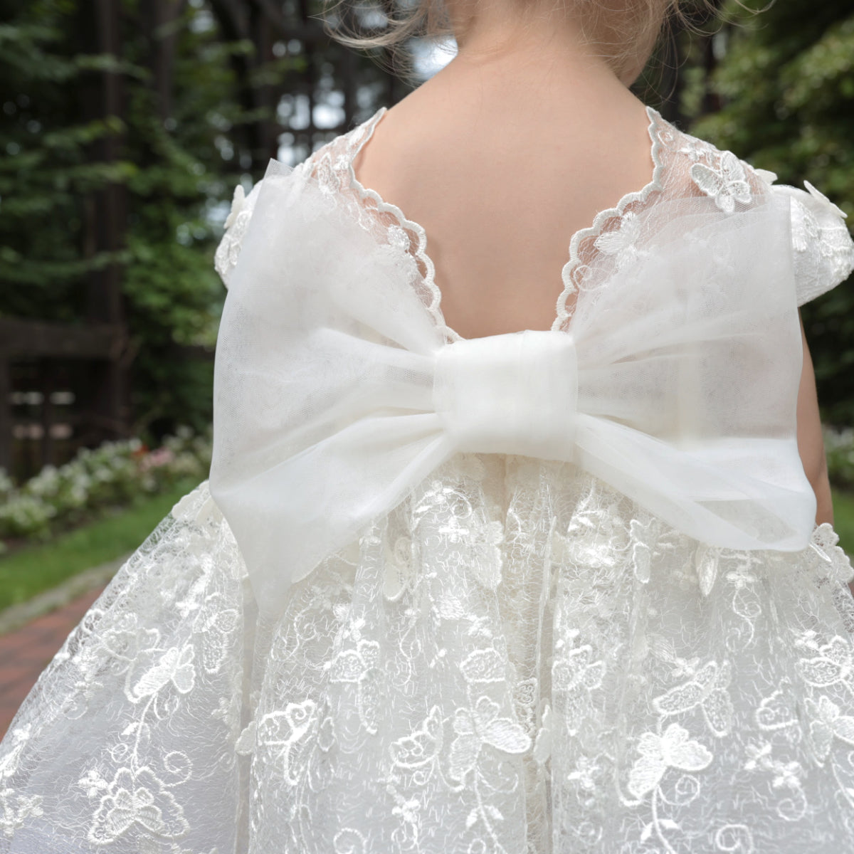 White lace dress with a large bow detail on the back, worn by a child, outdoors.