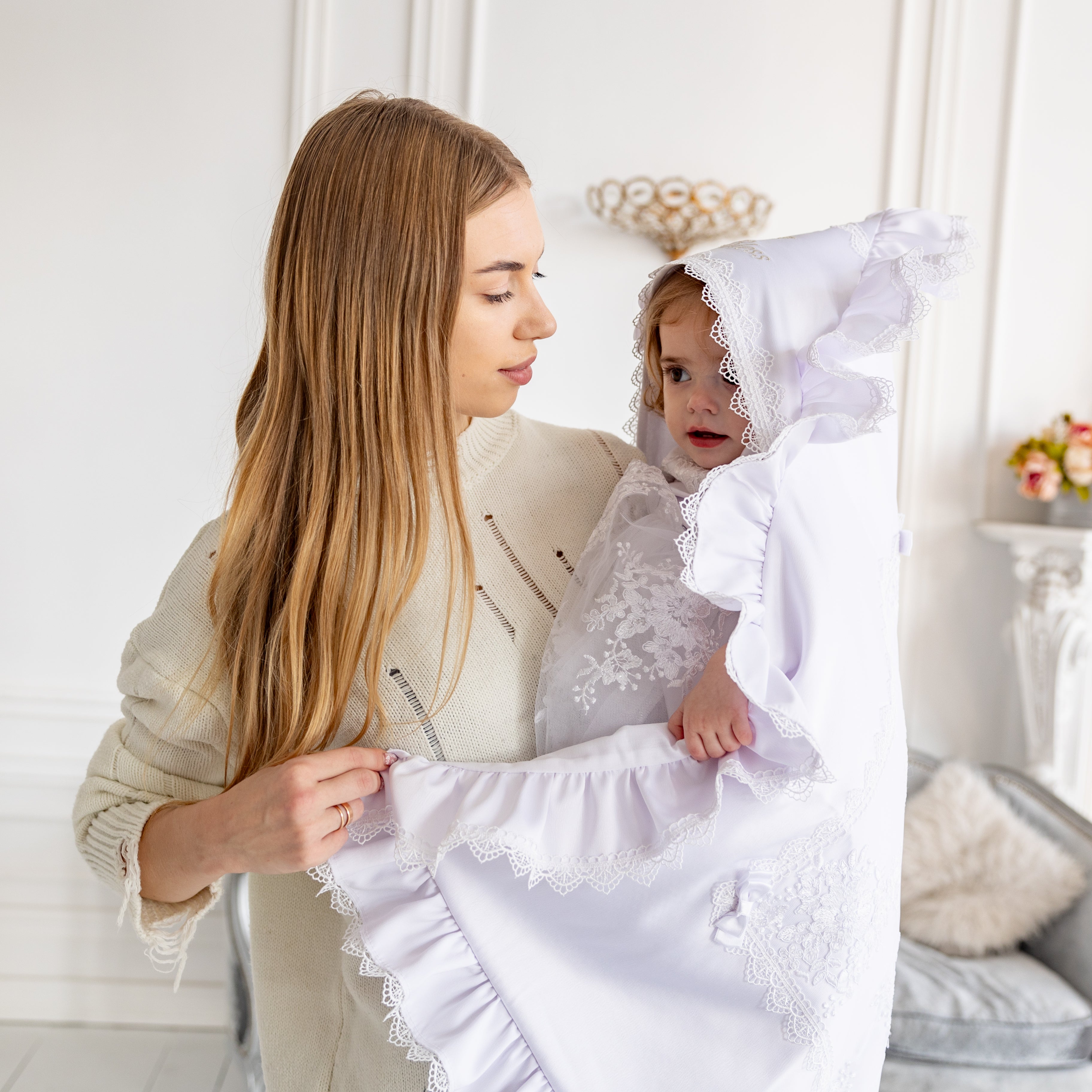 Woman holding a white lace baptism blanket with a child inside, in a softly lit room.
