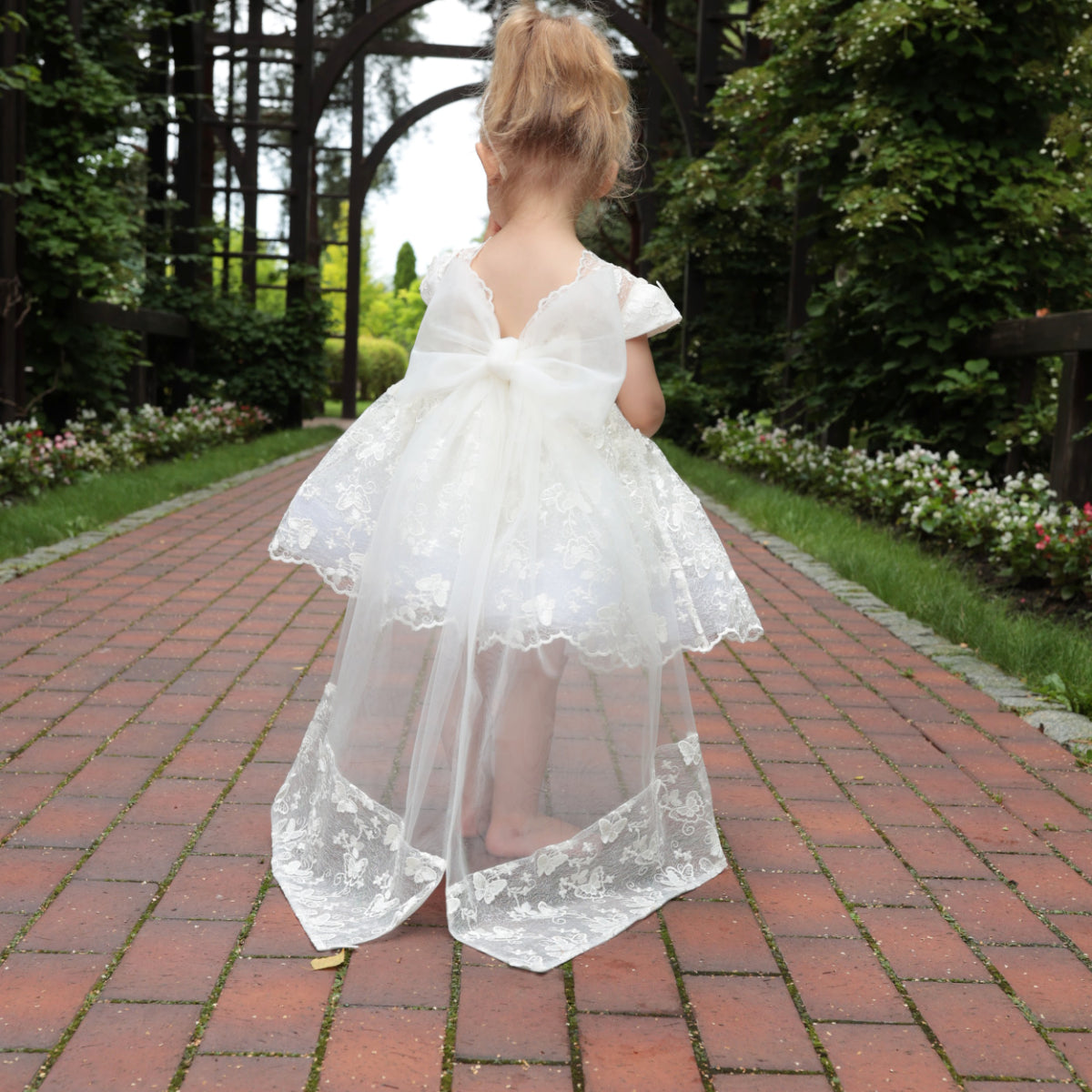 Young girl in a white lace dress walking on a brick path in a garden.