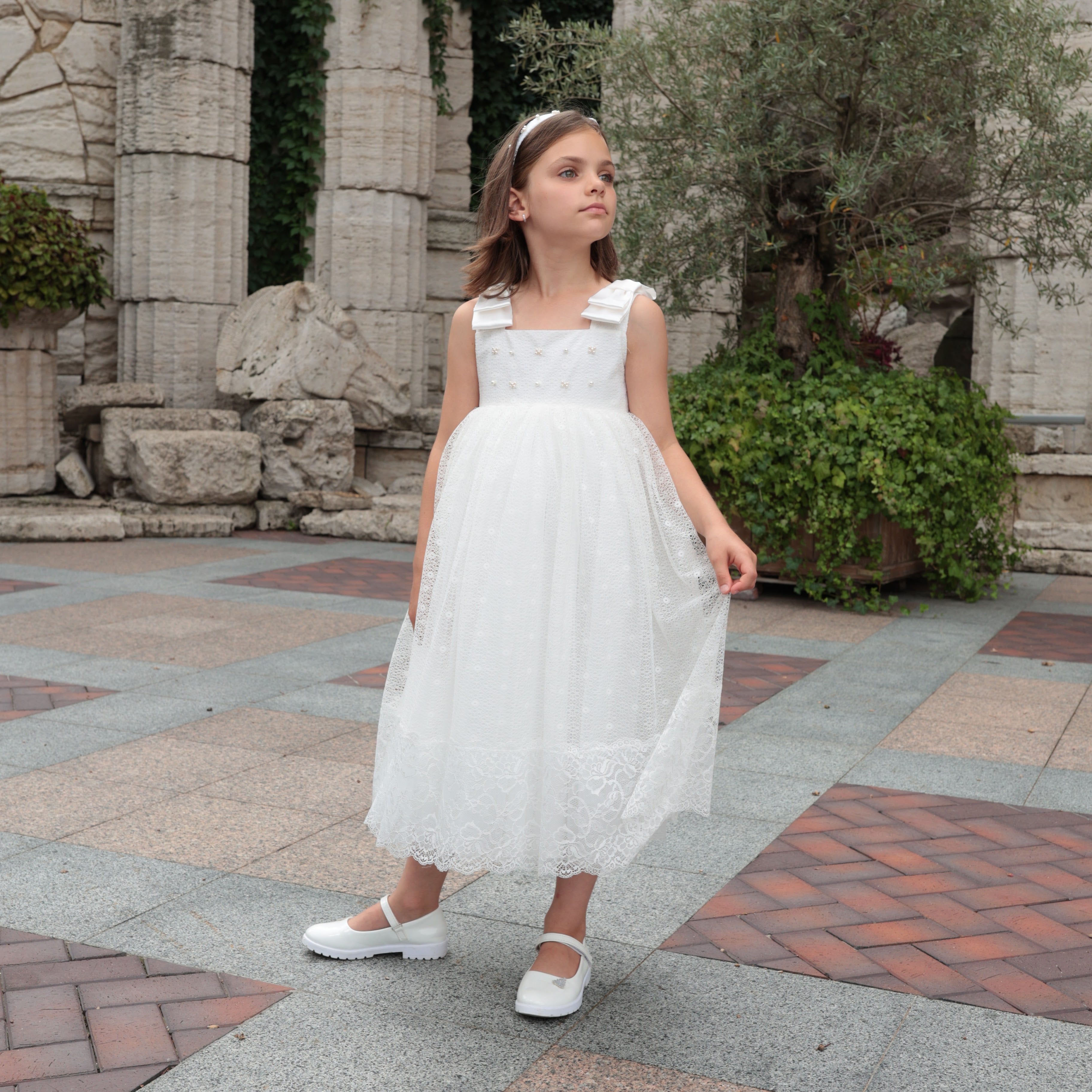 Young girl in a white dress standing on a stone pavement with greenery and stone columns in the background.