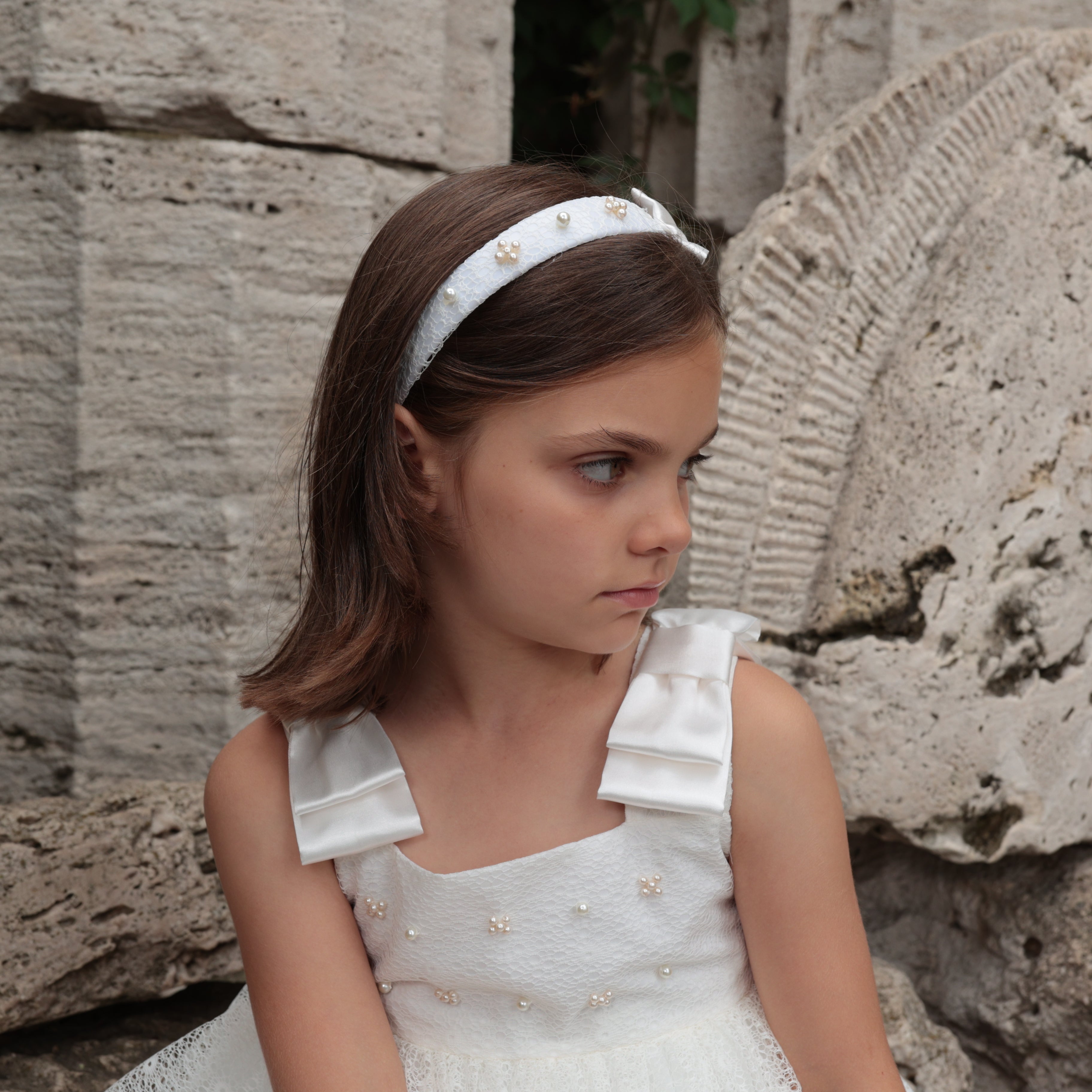 Young girl in a white dress with ruffled shoulders and a headband, sitting against a stone wall.