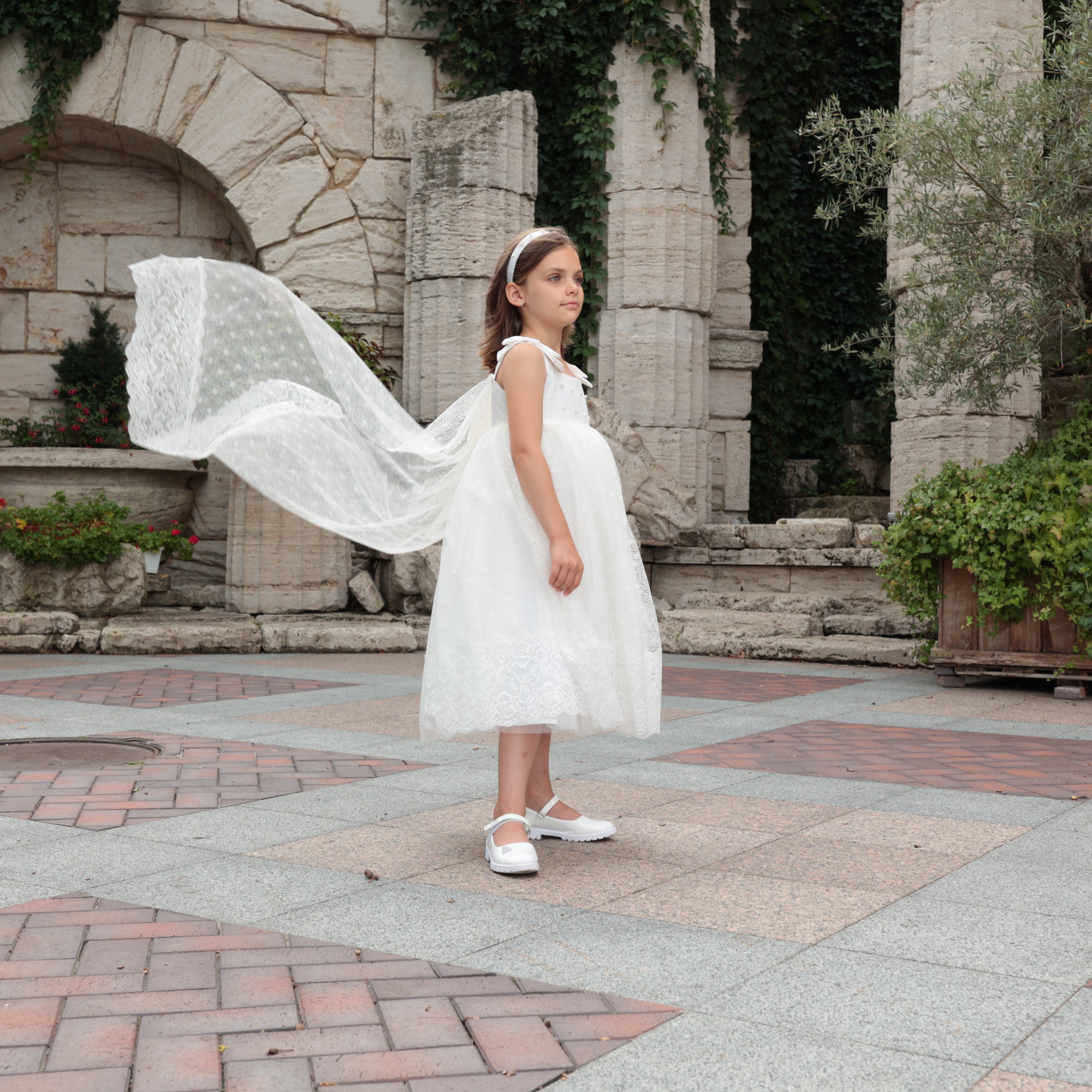 Young girl in a white dress standing in front of stone arches and plants.