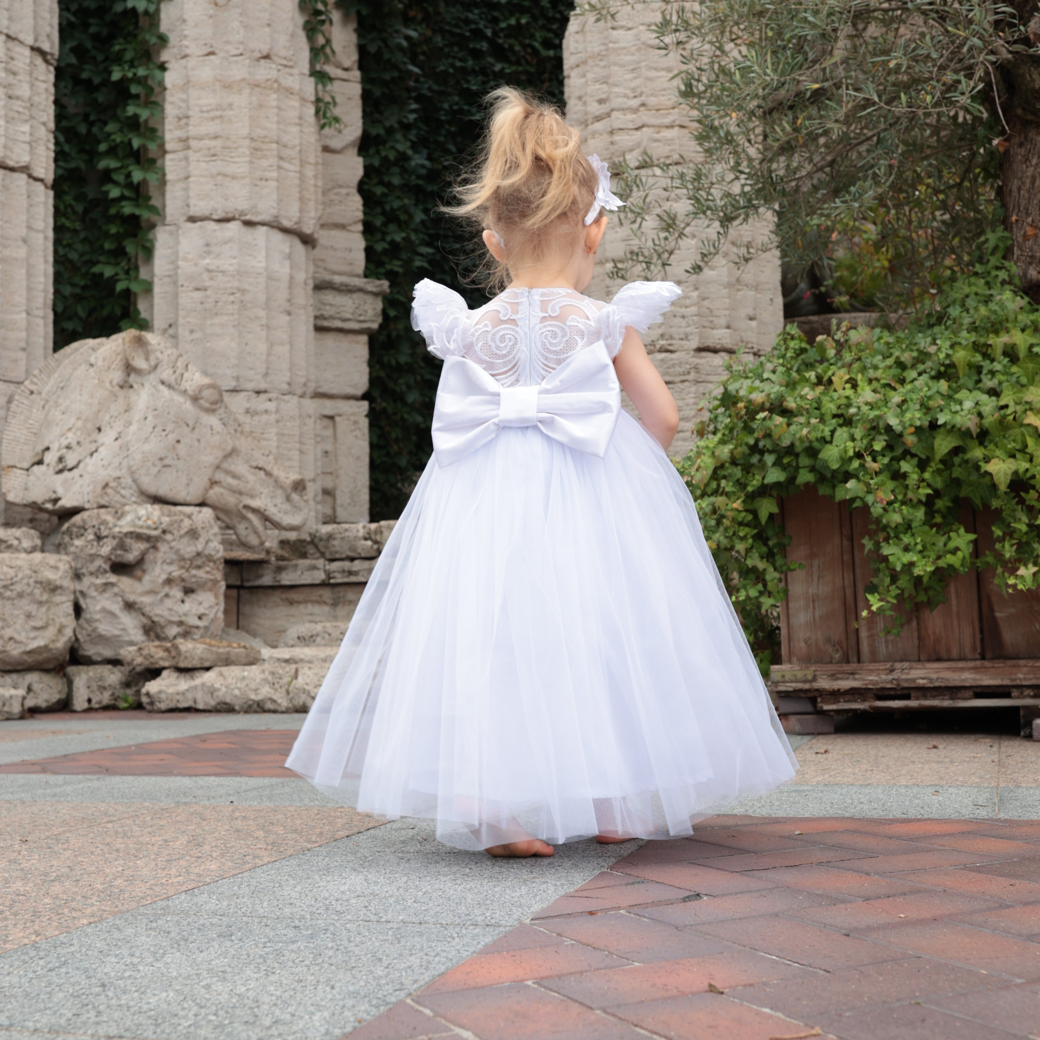 Young girl in a white dress standing in front of stone ruins and greenery.