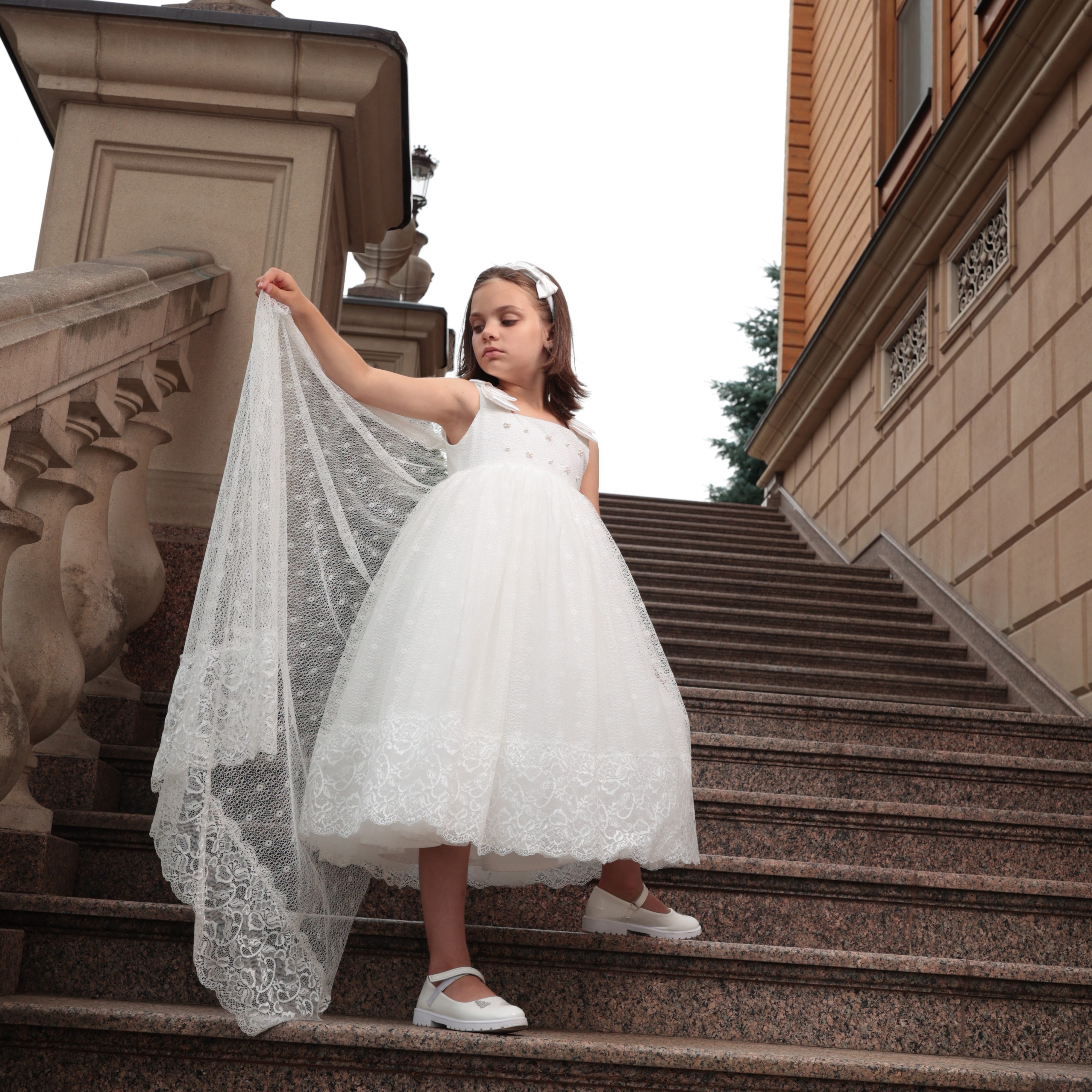 Young girl in ivory dress holding a lace veil on stone steps of an old building