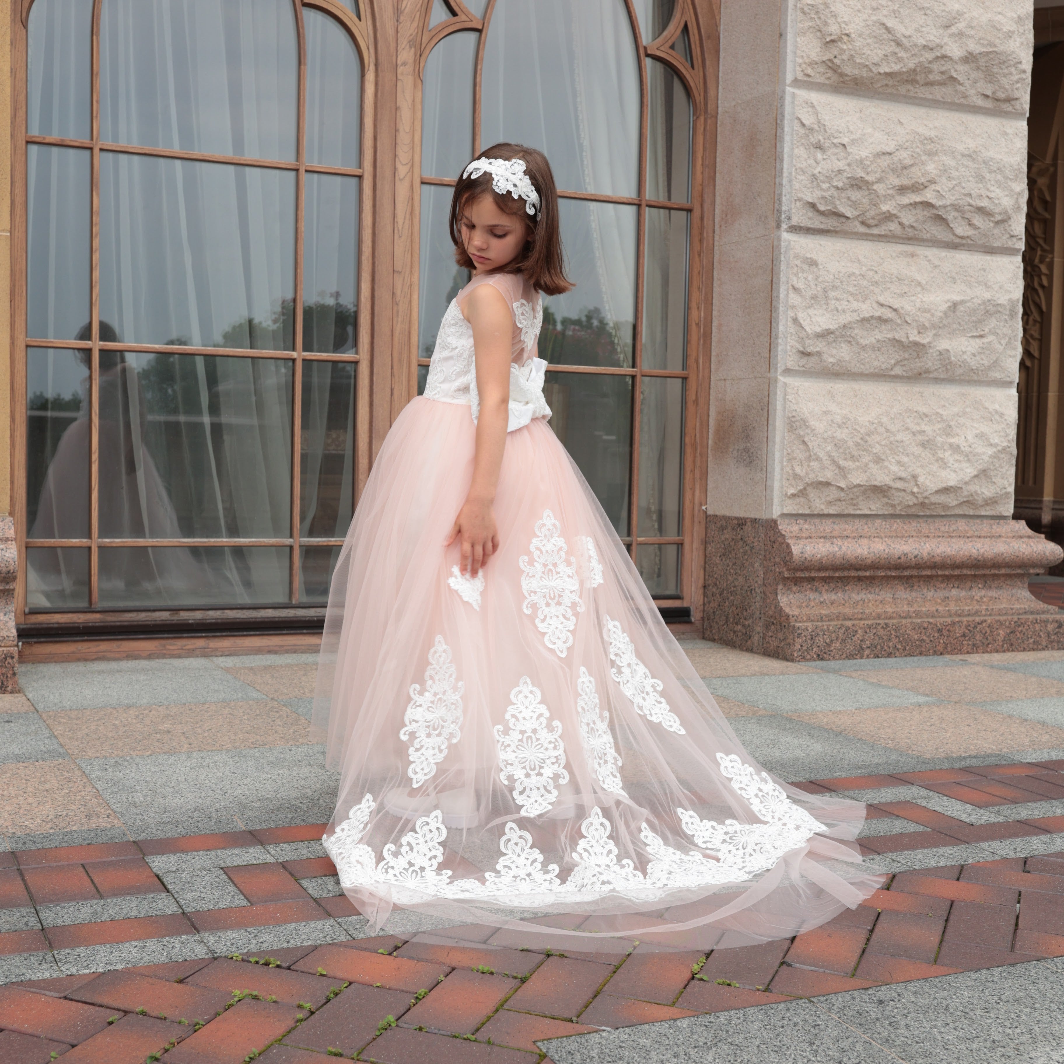 Young girl in a white top and pink floral dress standing in front of a stone building with large windows.