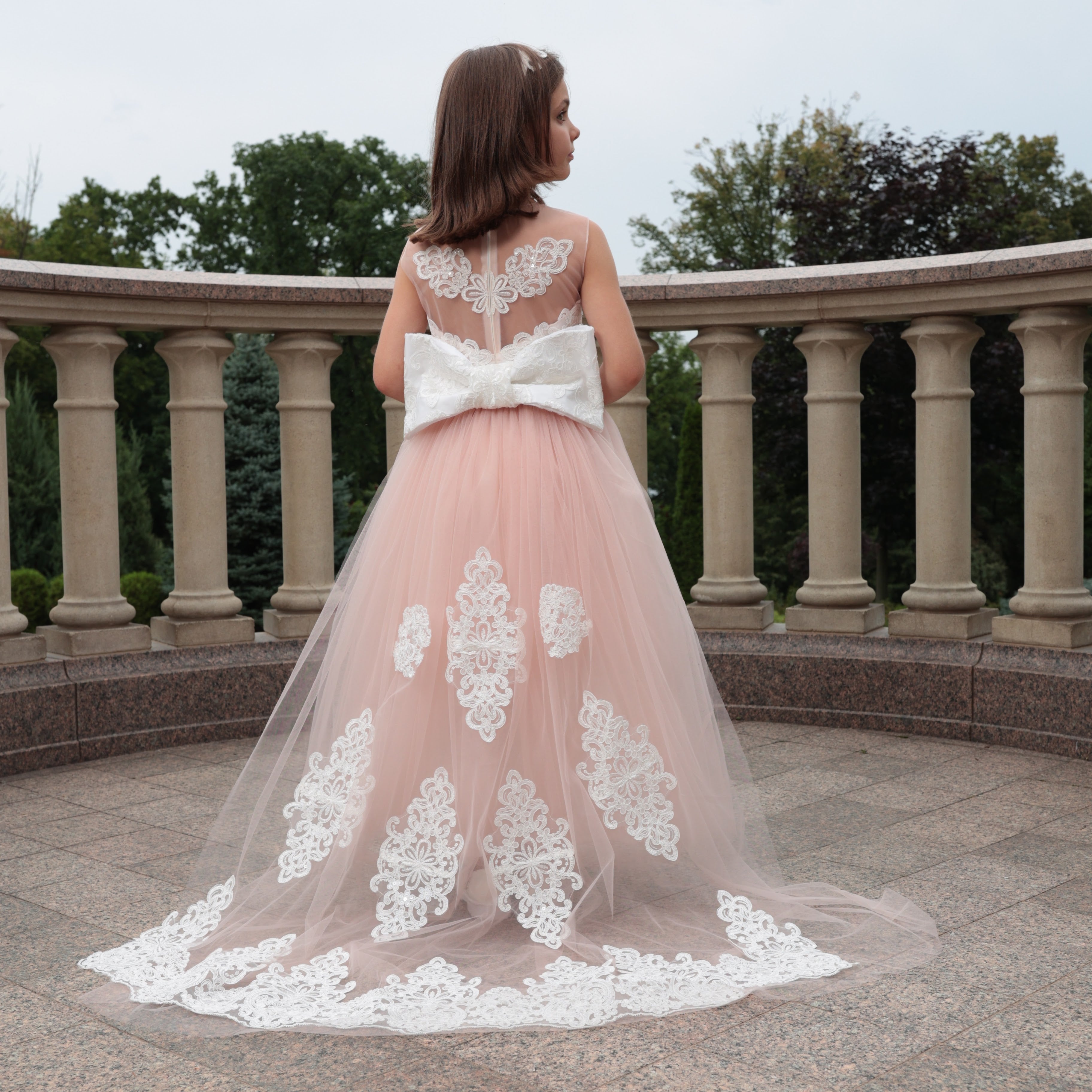 Giirl in a pink and white lace flower girl dress standing on a stone path with columns in the background.