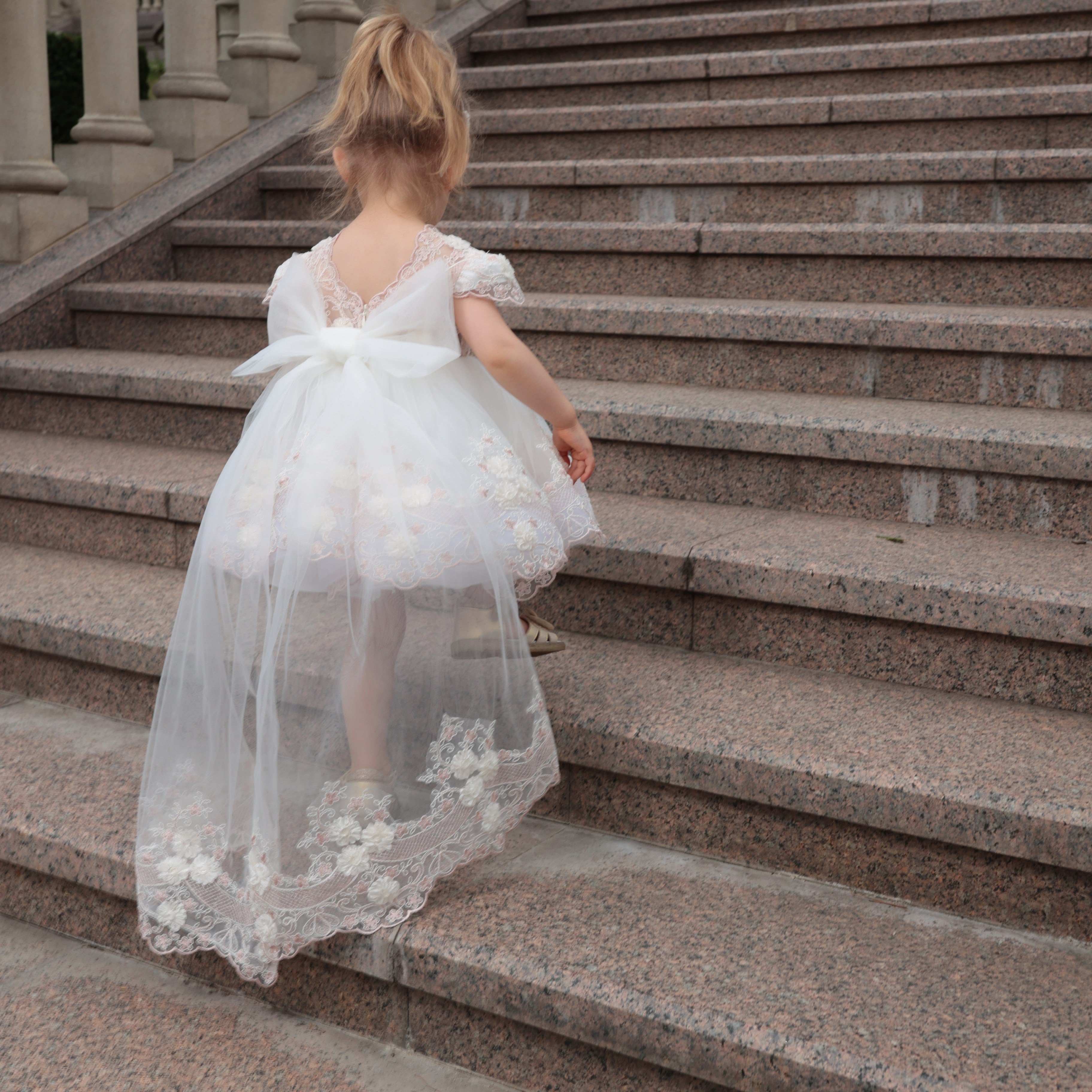 Child in a white dress with floral details walking up stone steps.