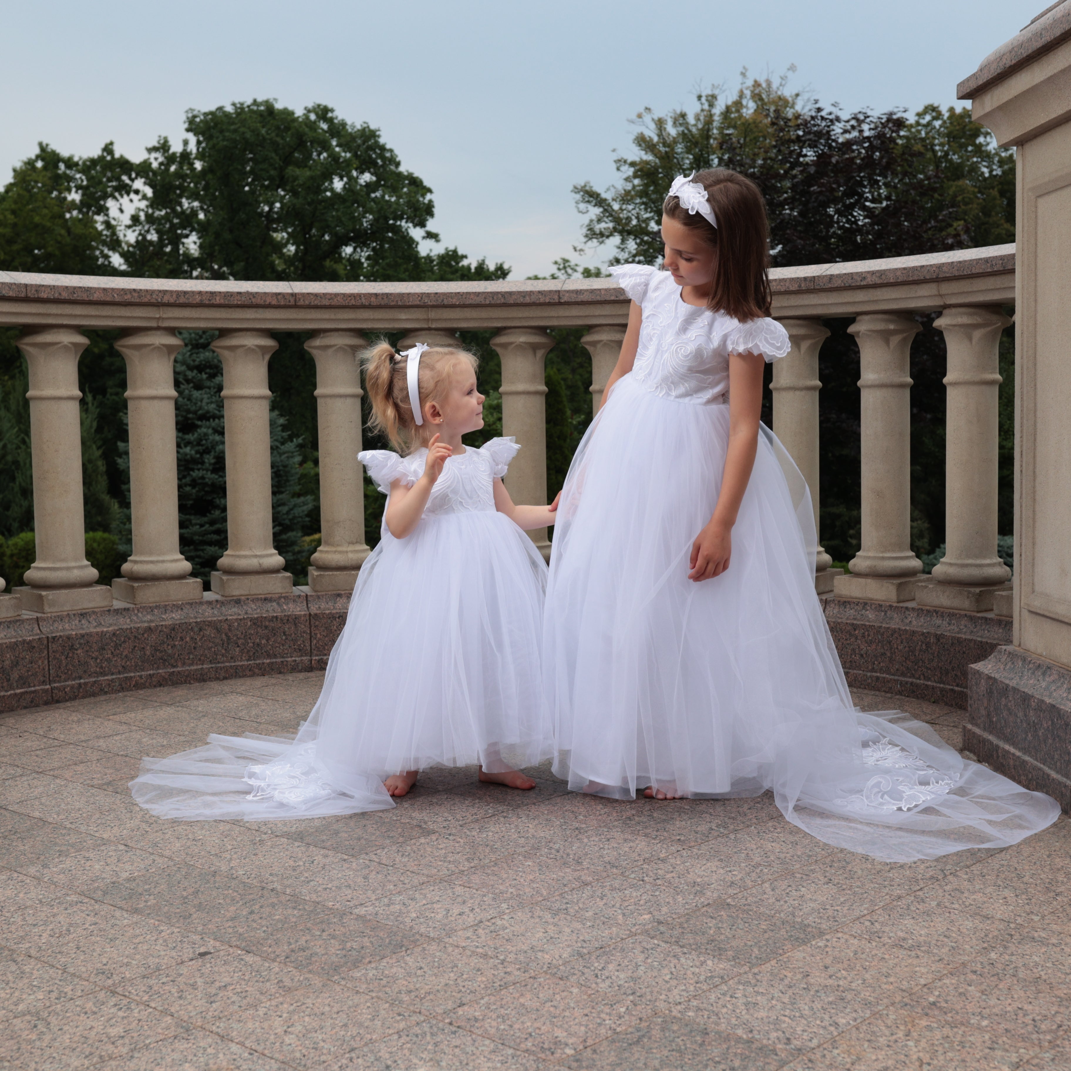 Two young girls in white dresses standing on a stone path with columns in the background.
