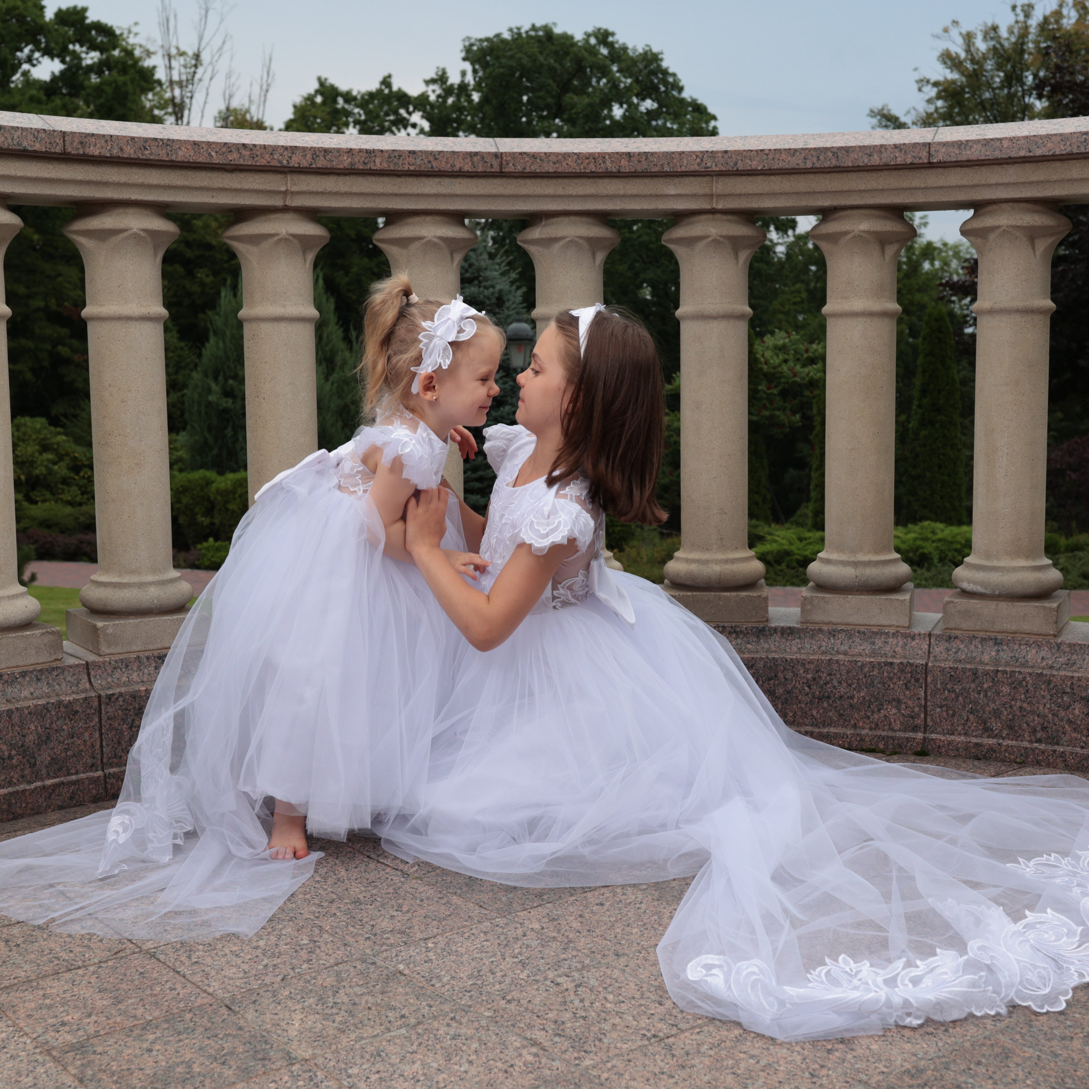 Two young girls in white dresses sitting on a stone ledge with columns in the background.