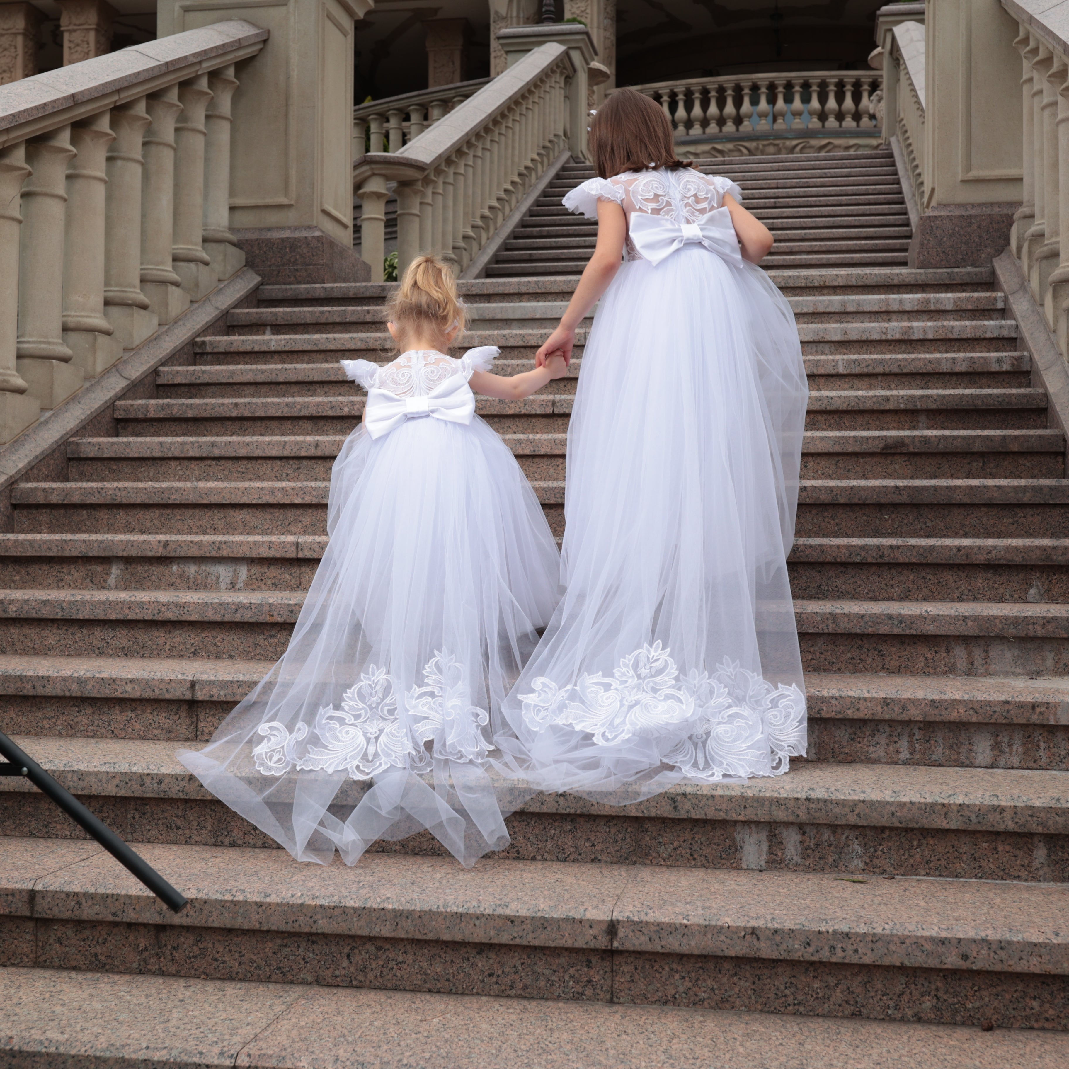 Two women in white dresses with floral details walking down a staircase.