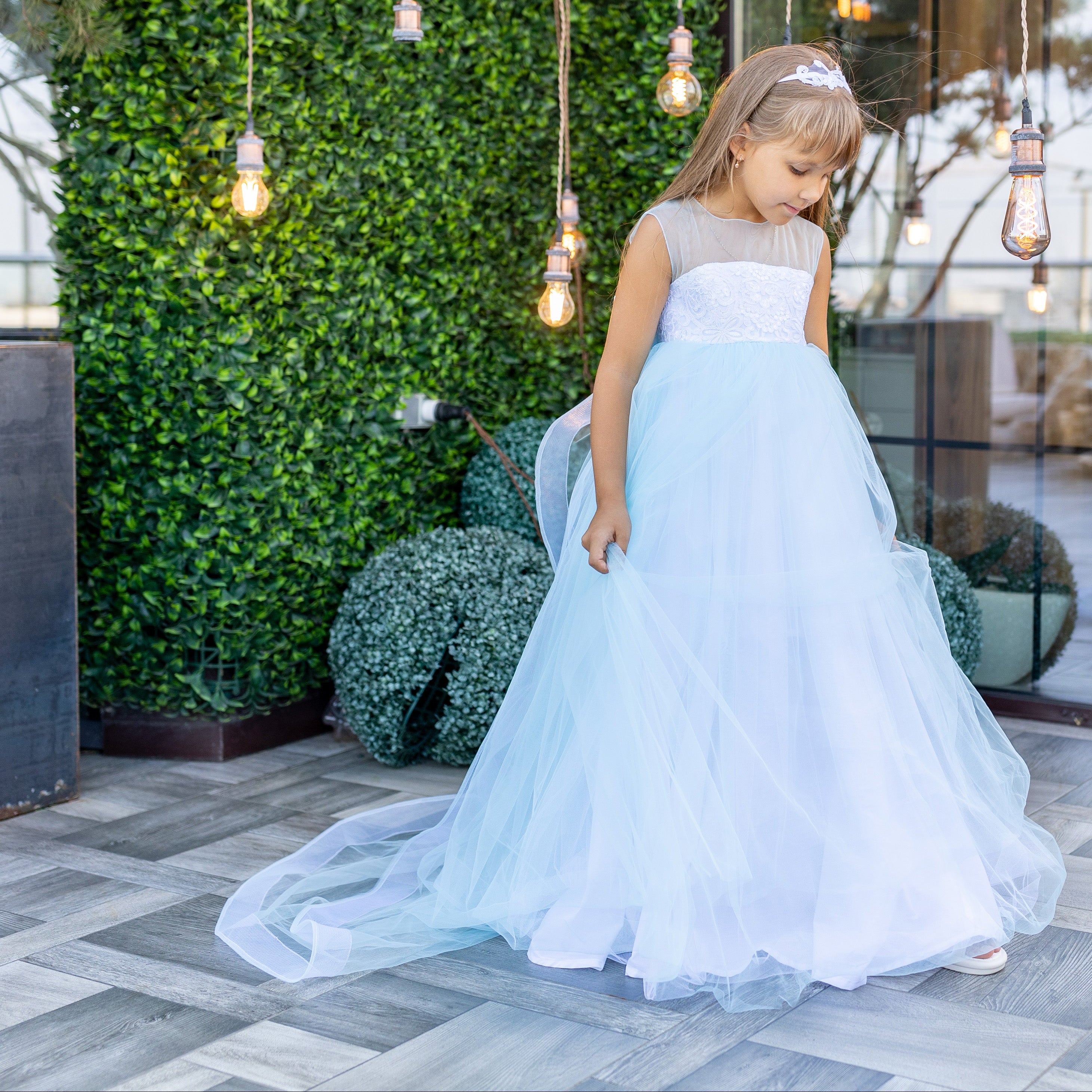 Young girl in a light blue dress standing in an outdoor setting with greenery and hanging lights.