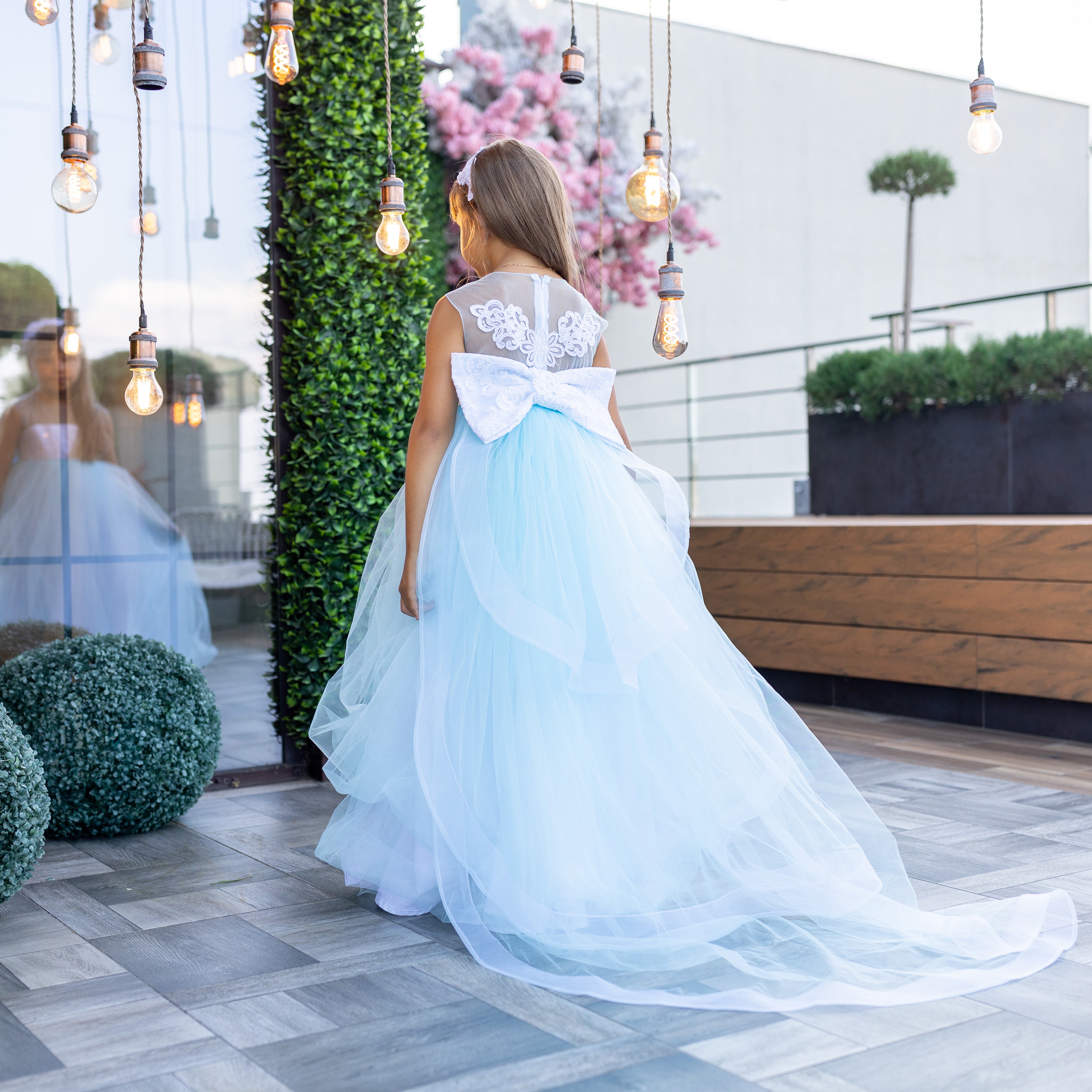 Woman in a light blue dress with a large white bow standing on a rooftop patio.