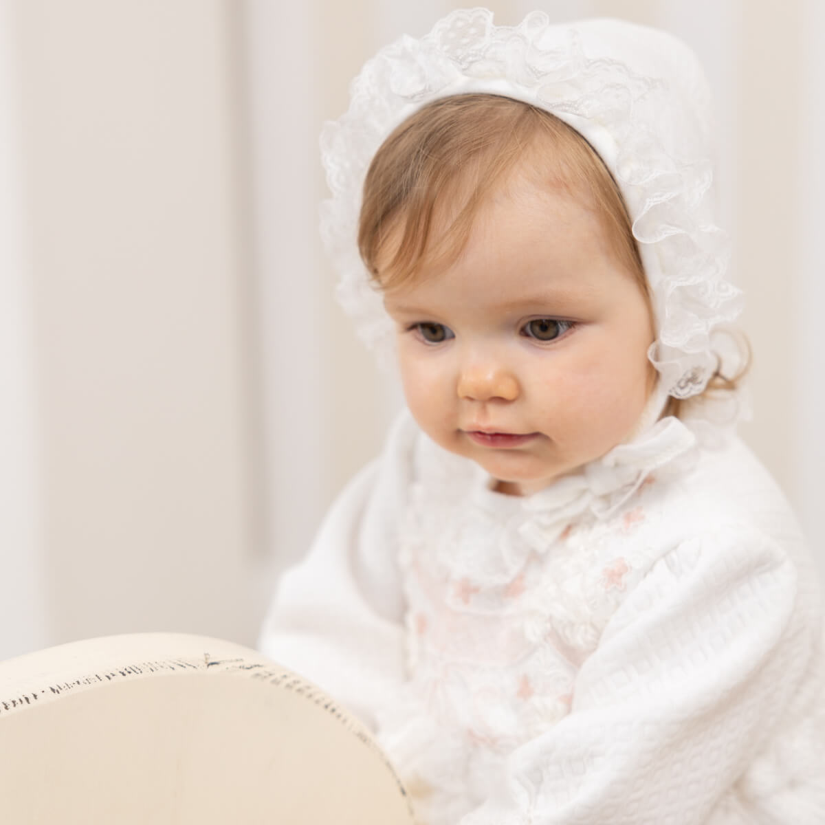 Baby wearing a Lace Baptism Bonnet and white outfit against a neutral background