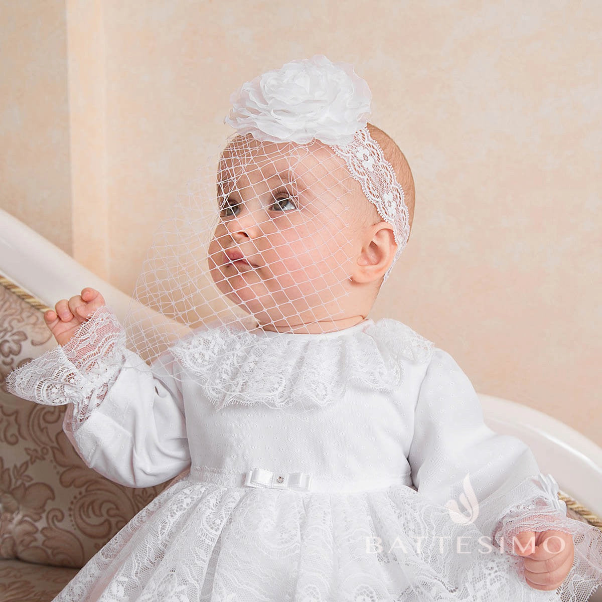 Girl in a white lace dress with the baby headband and veil, sitting on a decorative chair.