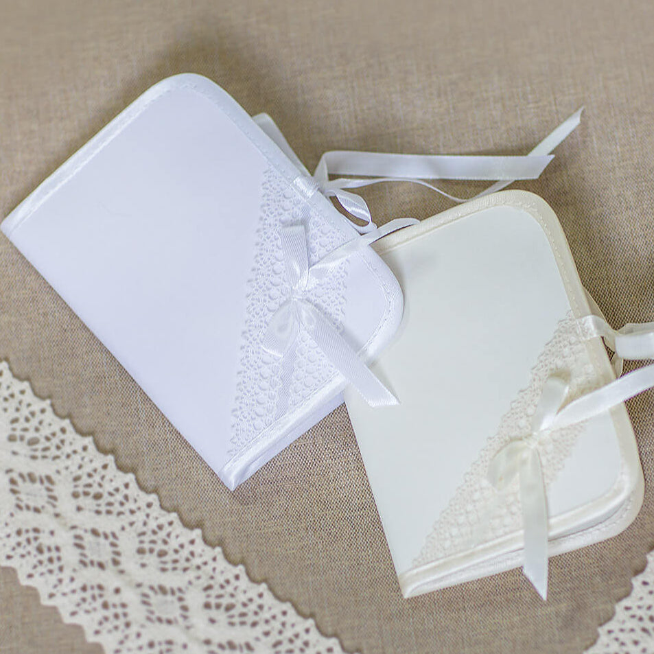 Two small white satin-covered booklets or cards with lace trim and ribbon ties, arranged on a beige fabric background with white lace trim.