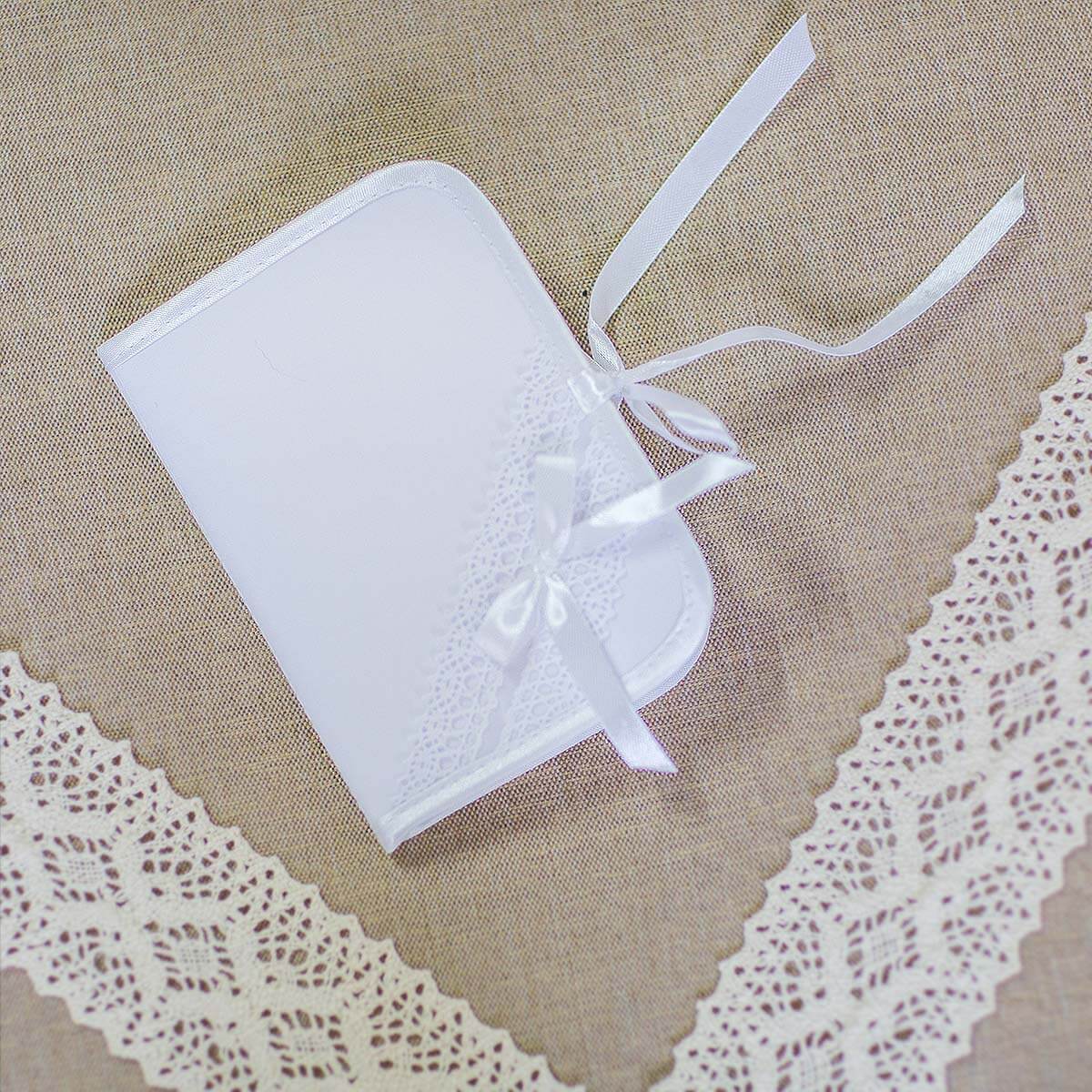 A small white rectangular gift box or keepsake case with rounded corners, decorated with a white lace panel and tied with a white satin ribbon, placed on a beige fabric background with white lace trim.