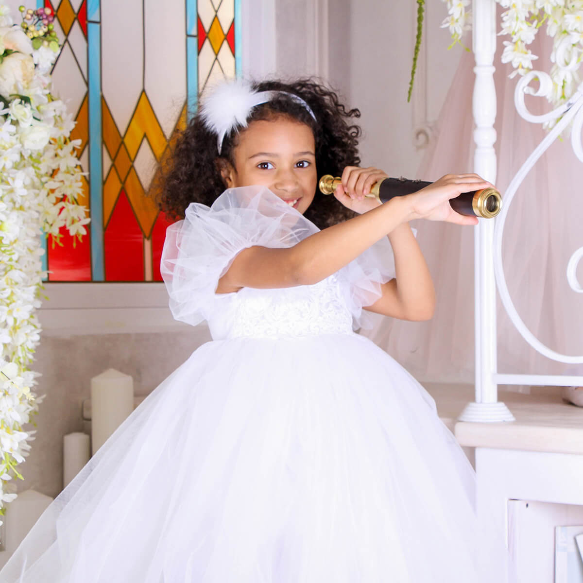Young girl in a white dress holding a golden scepter in front of a stained glass window.
