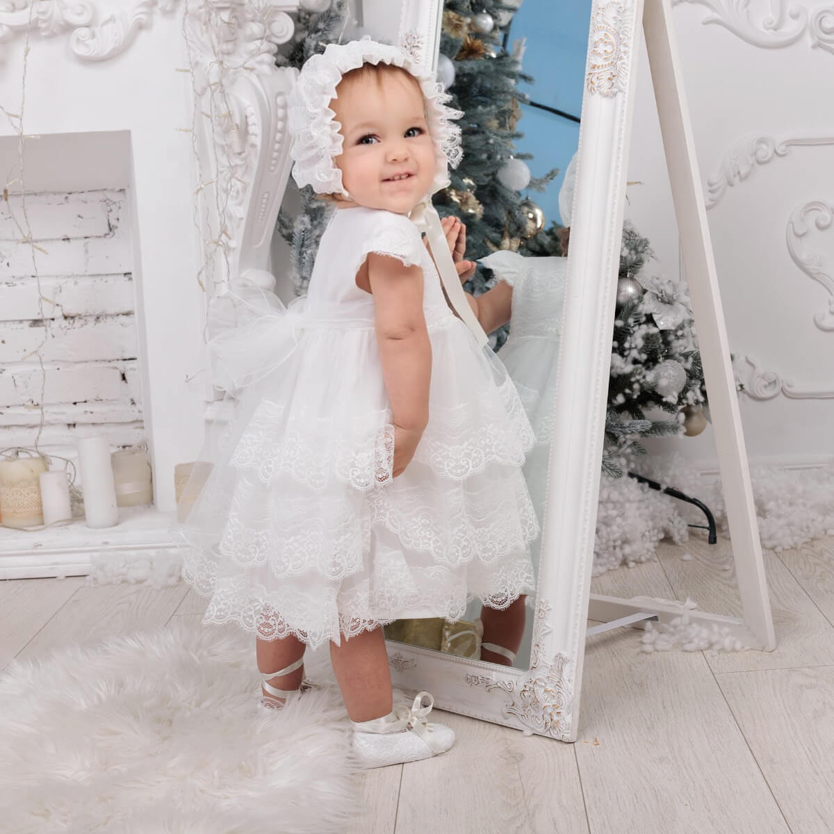 Baby in a Lace Baptism Bonnet standing in front of a mirror with a Christmas tree in the background.
