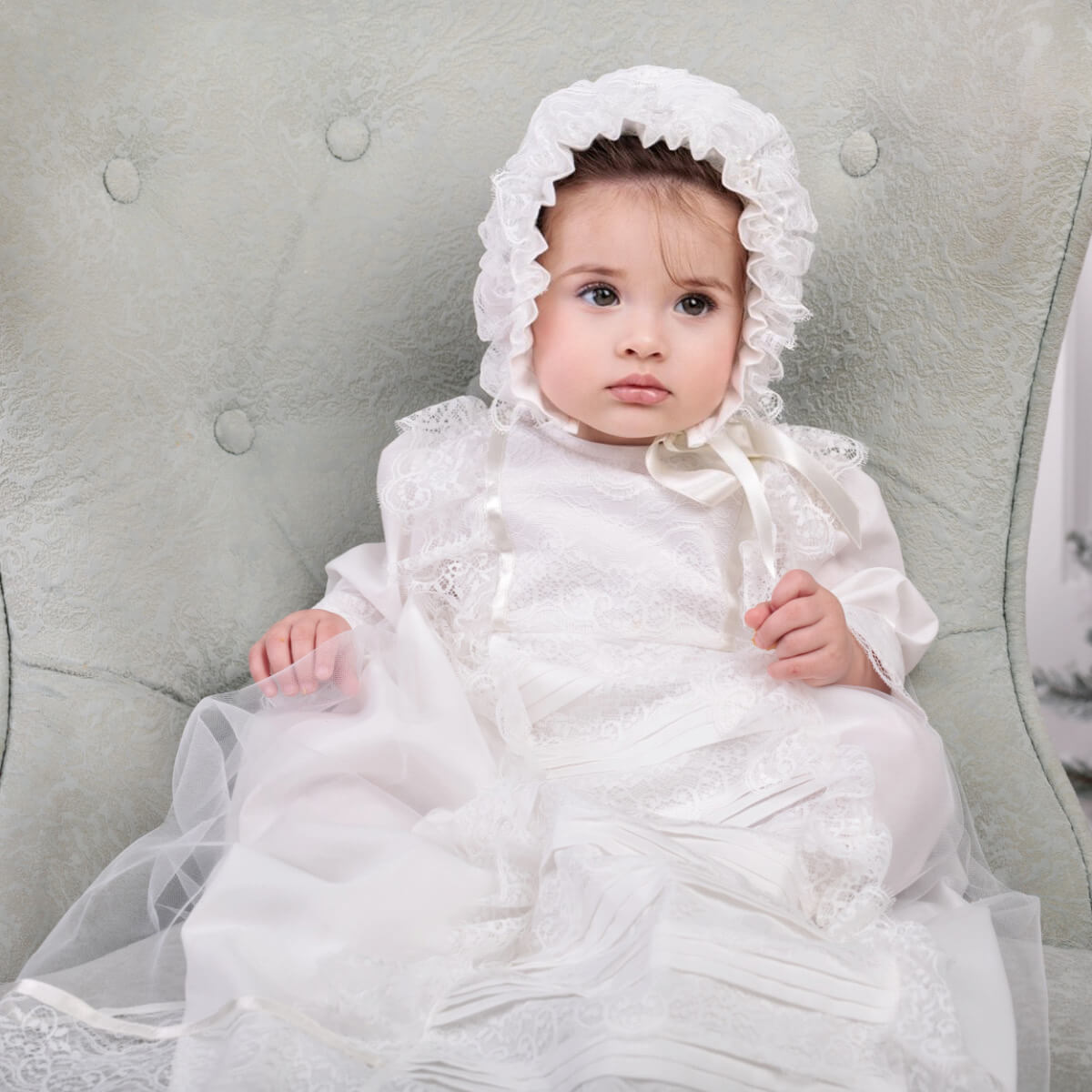 Baby in a white lace dress and Lace Baptism Bonnet sitting on a gray cushion.