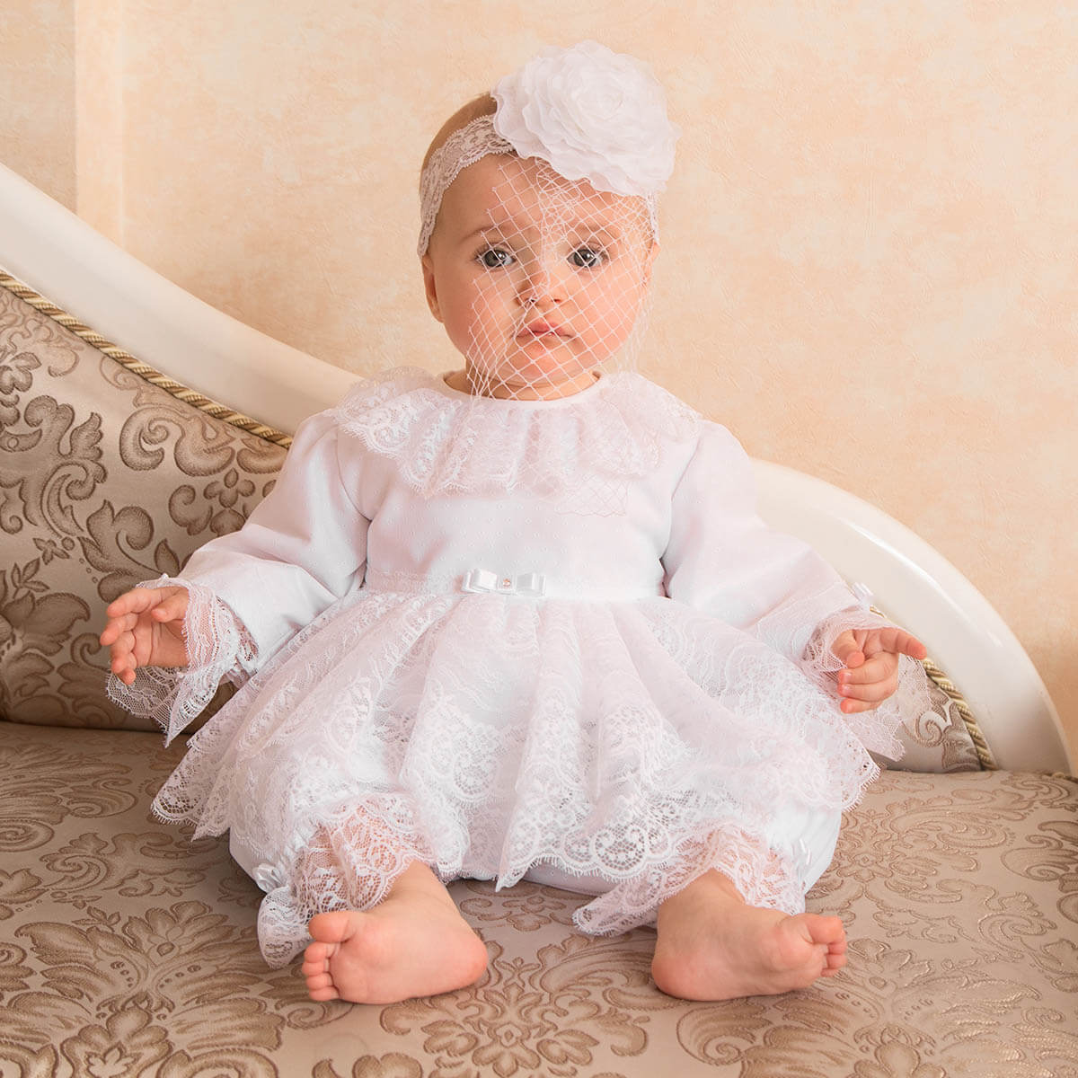 Baby in a white lace romper sitting on a patterned couch.