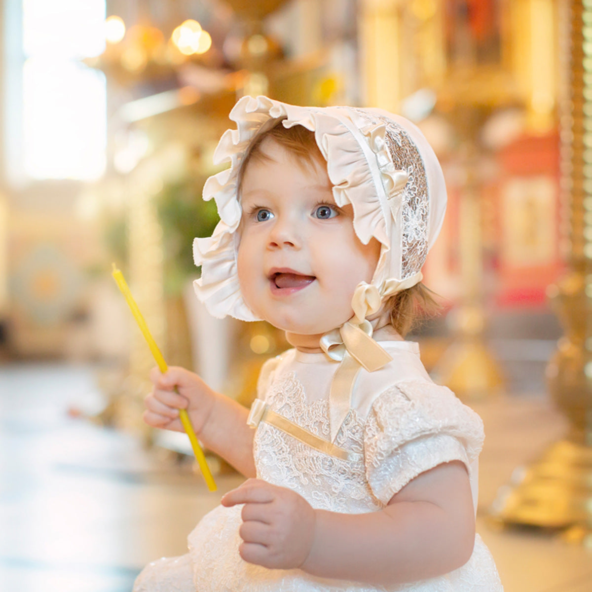 Child wearing a Silk Christening Bonnets and holding a yellow wand in front of a blurred carousel background