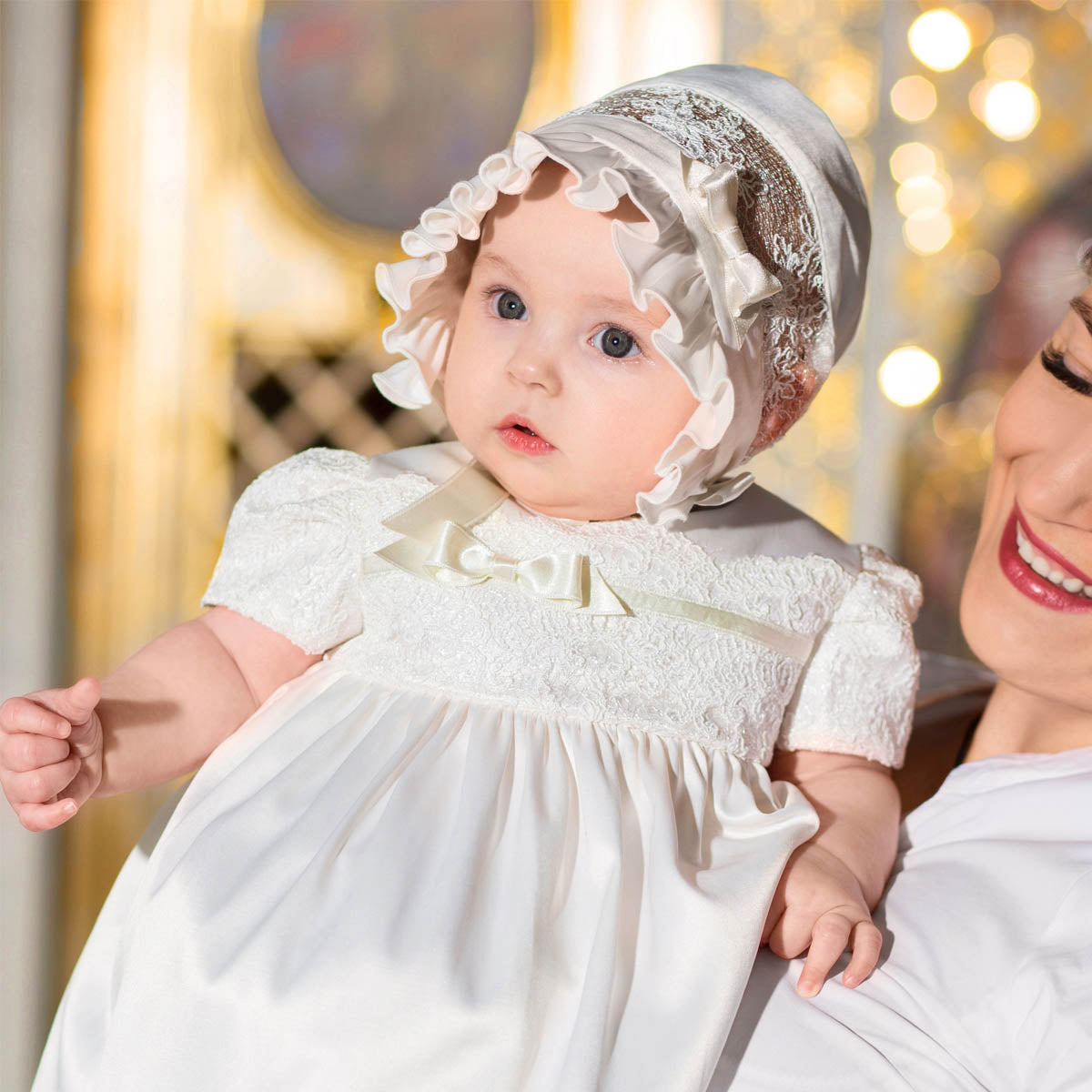 Baby in a white dress and bonnet held by an adult against a blurred background