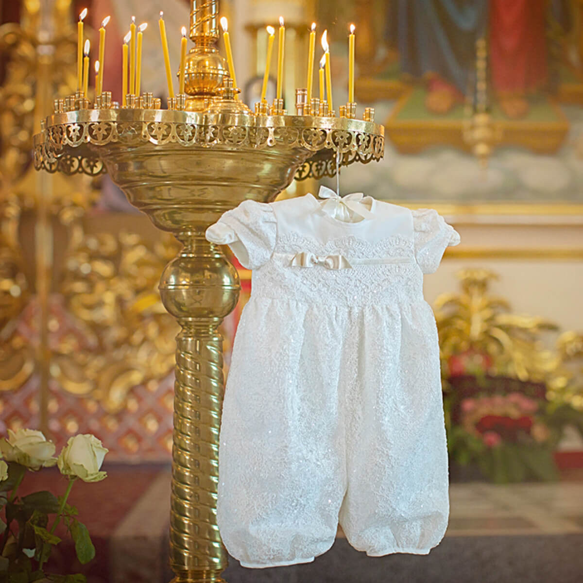 White baptism bubble romper hanging from a gold candle holder in a decorated room.