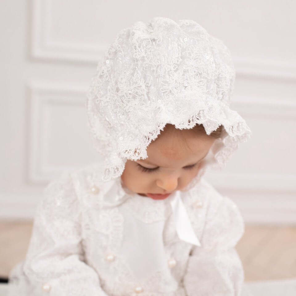 Baby wearing a white lace bonnet and tGirls Christening Gown against a neutral background