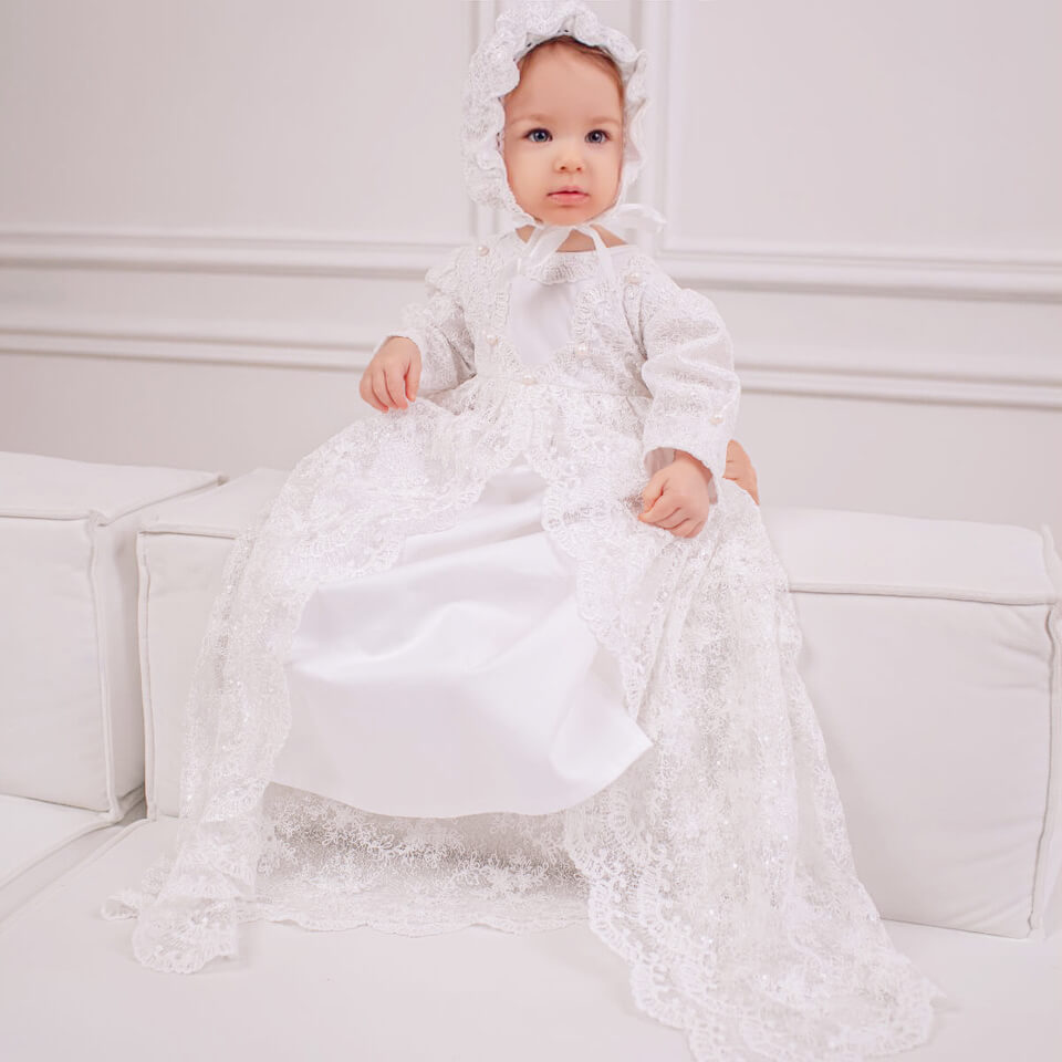 Baby in a Girls Christening Gown sitting on a white couch.