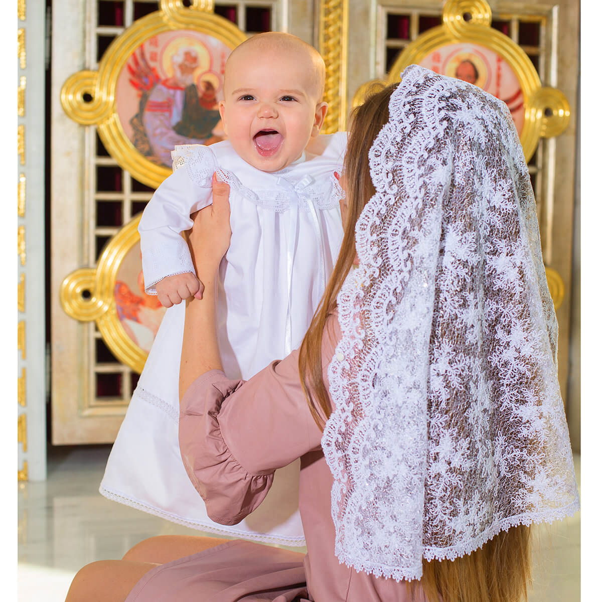 Woman holding a baby in a white dress with a Church Mantilla Veil, in front of religious iconography.