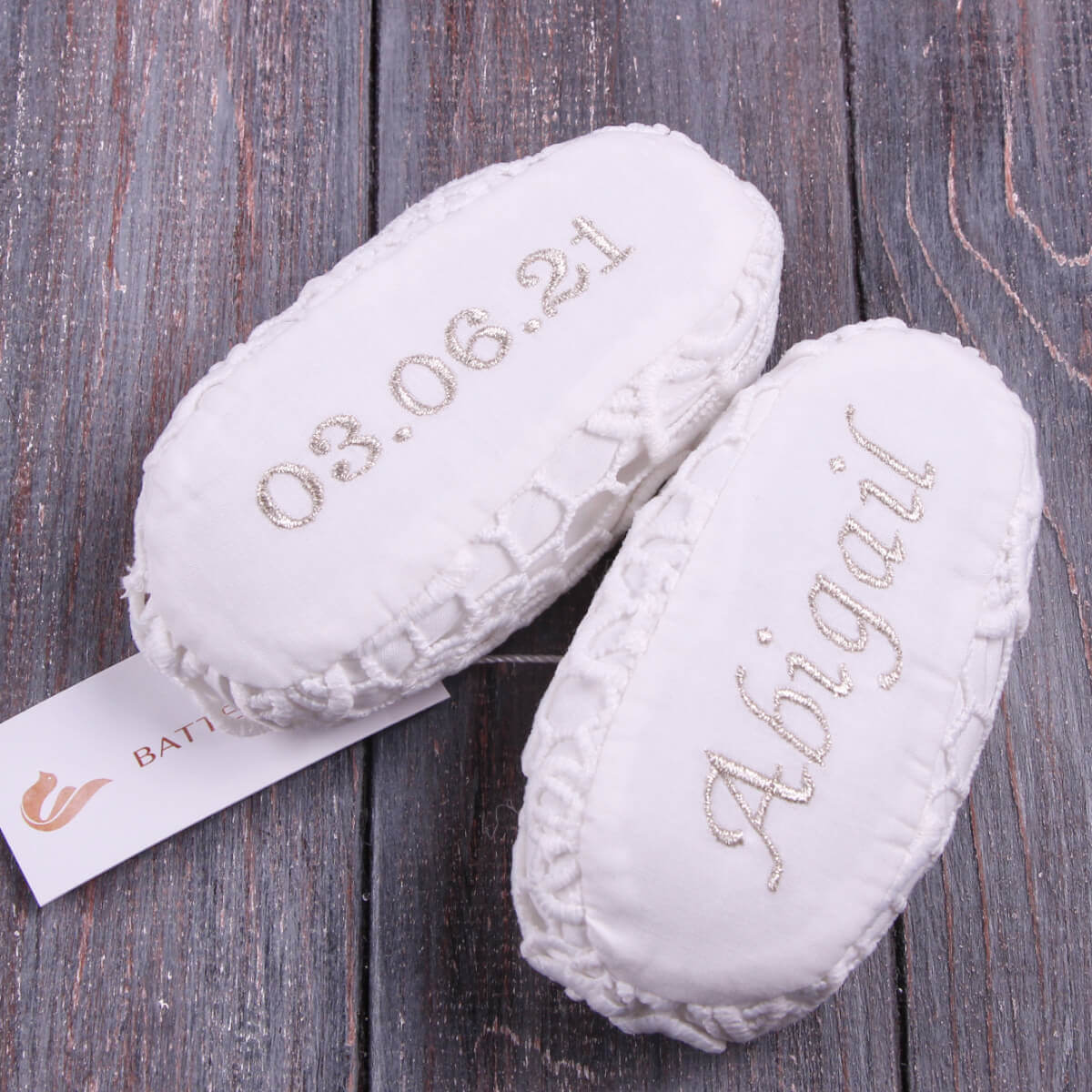 White baby booties with embroidered text on a wooden surface