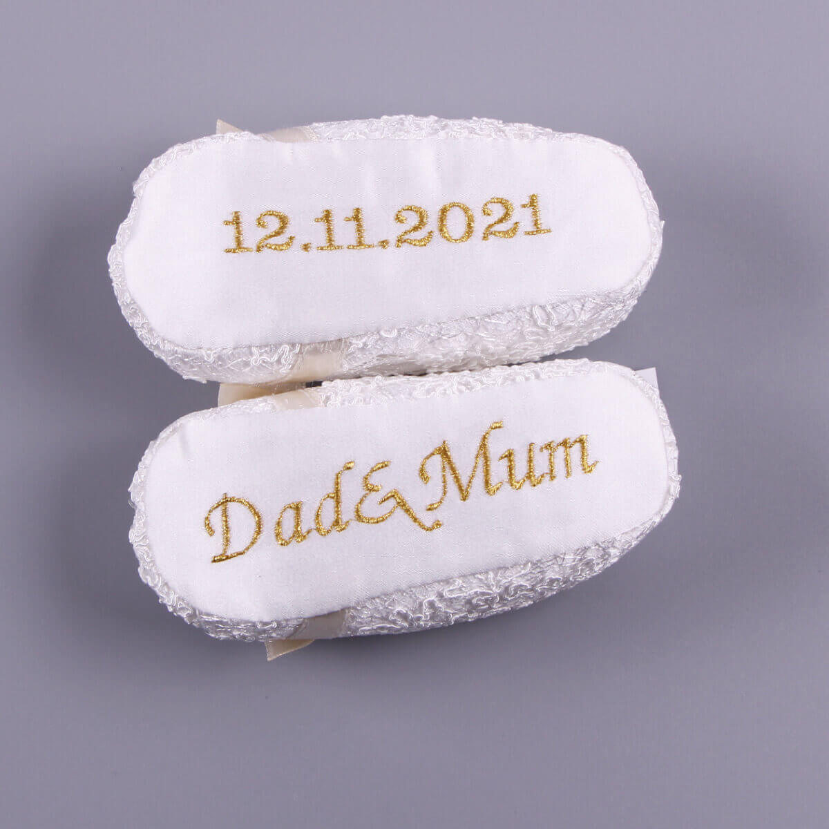 Two white embroidered White baptism booties with '12.11.2021' and 'Dad & Mum' text on a gray background.