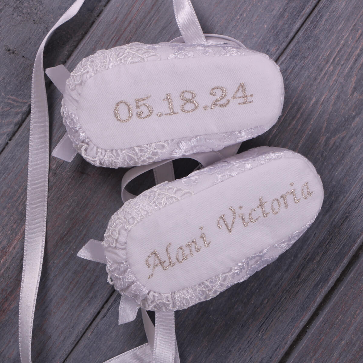 White baptism booties with '05.18.24' and 'Alani Victoria' embroidery on a wooden surface