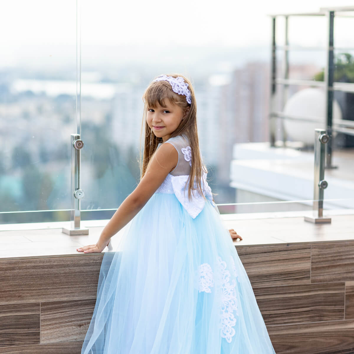 Young girl in a light blue dress with white floral details standing on a balcony.