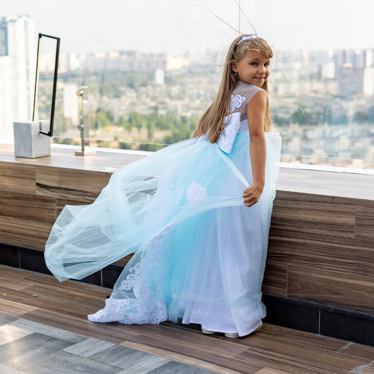 Young girl in a light blue dress standing on a rooftop with a cityscape view.