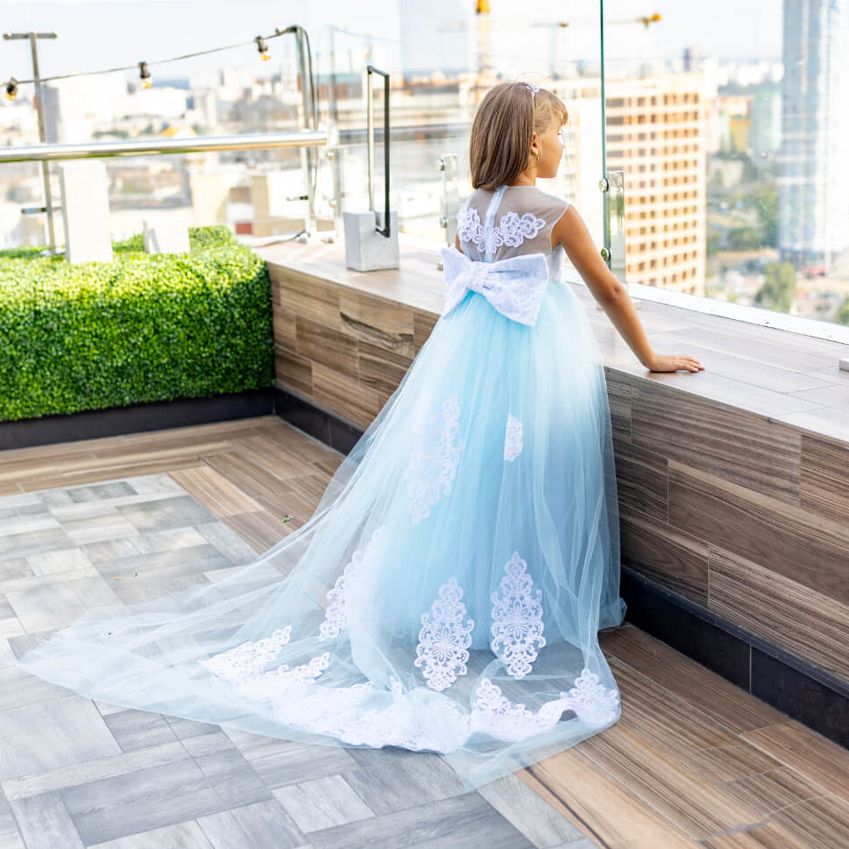 Woman in a light blue dress with white lace details standing on a rooftop terrace with cityscape view.