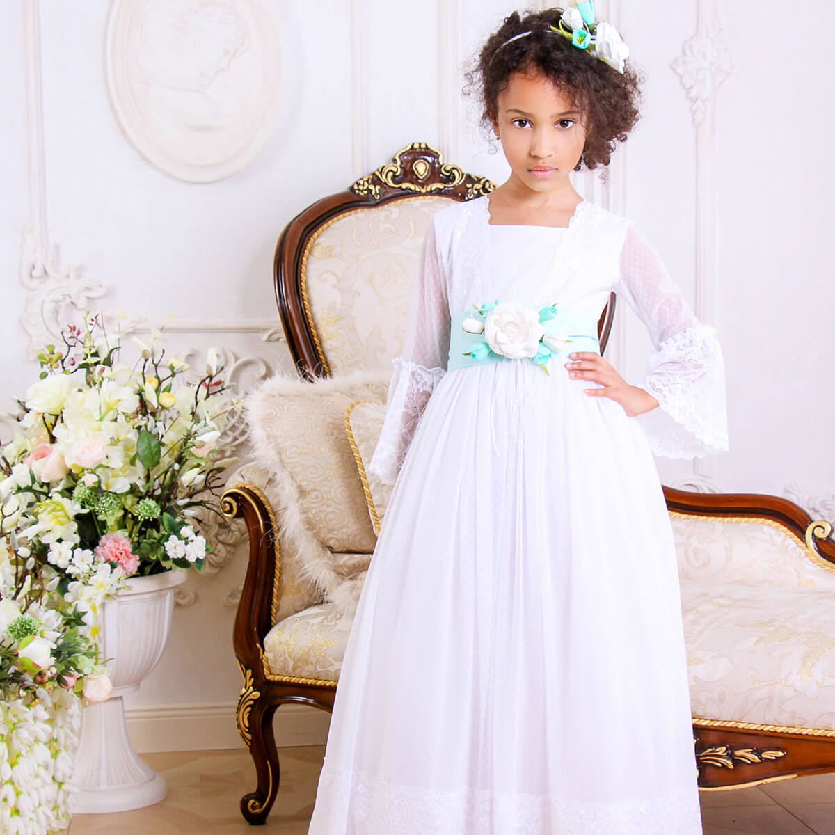 Young girl in a Traditional Communion Dress with floral decorations sitting on an ornate chair in a decorated room.