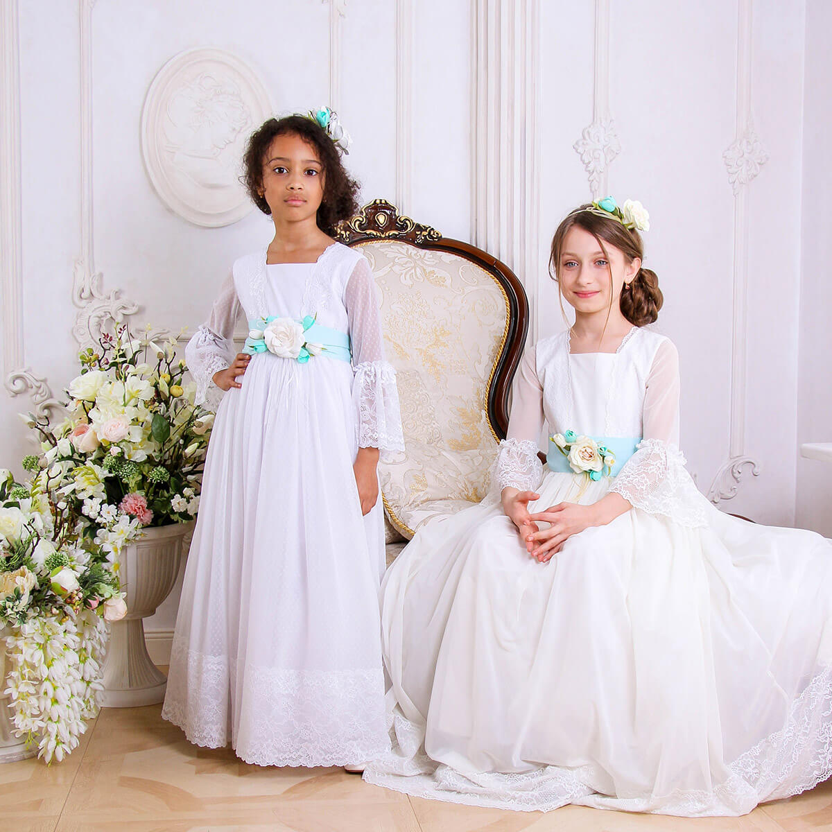 Two young girls in white dresses sitting on a decorative chair with floral arrangements around.